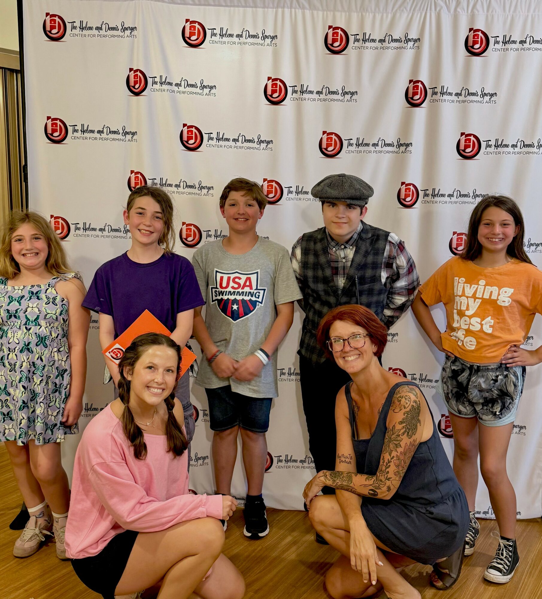 Six children and one adult woman pose in front of a backdrop with logos, smiling and standing or sitting together.