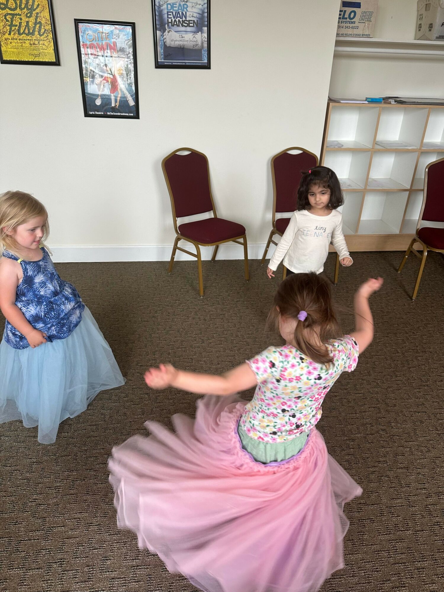 Two children playing indoors, one girl in a pink tutu and another girl in a blue dress, with chairs and framed pictures in the background.
