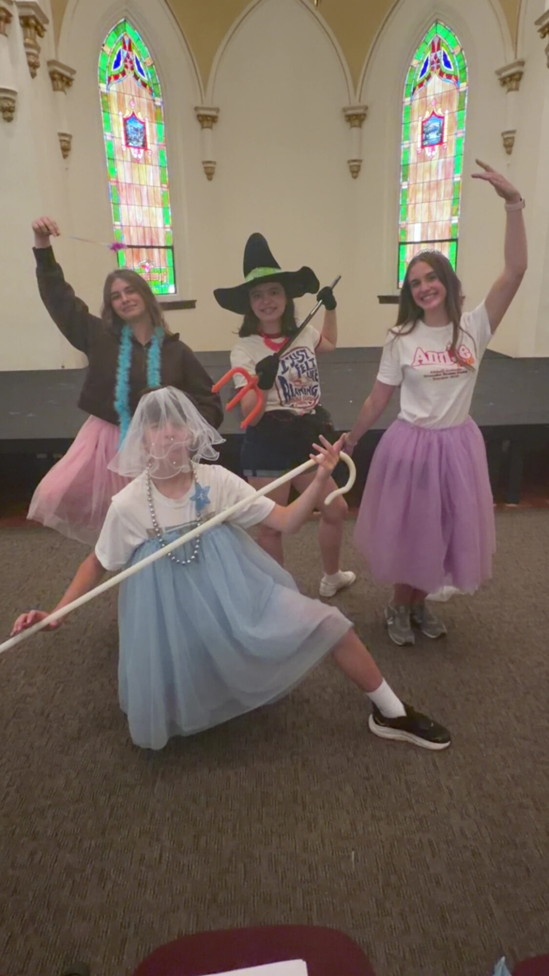 Four children in costumes pose inside a church with stained glass windows, one dressed as a witch, others in colorful skirts.