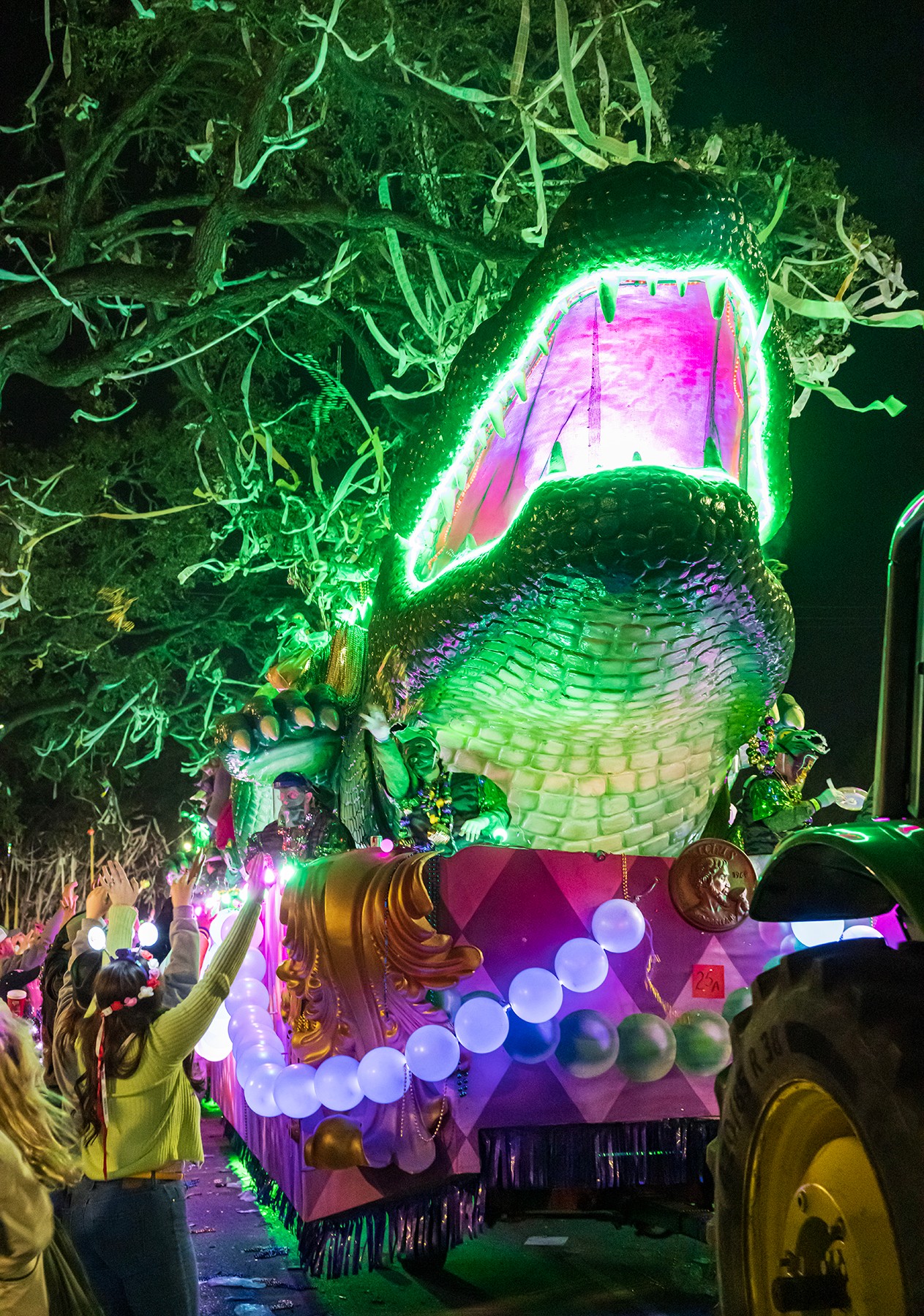 Colorful parade float with a large open-mouthed crocodile head, illuminated with bright lights, surrounded by people at night.