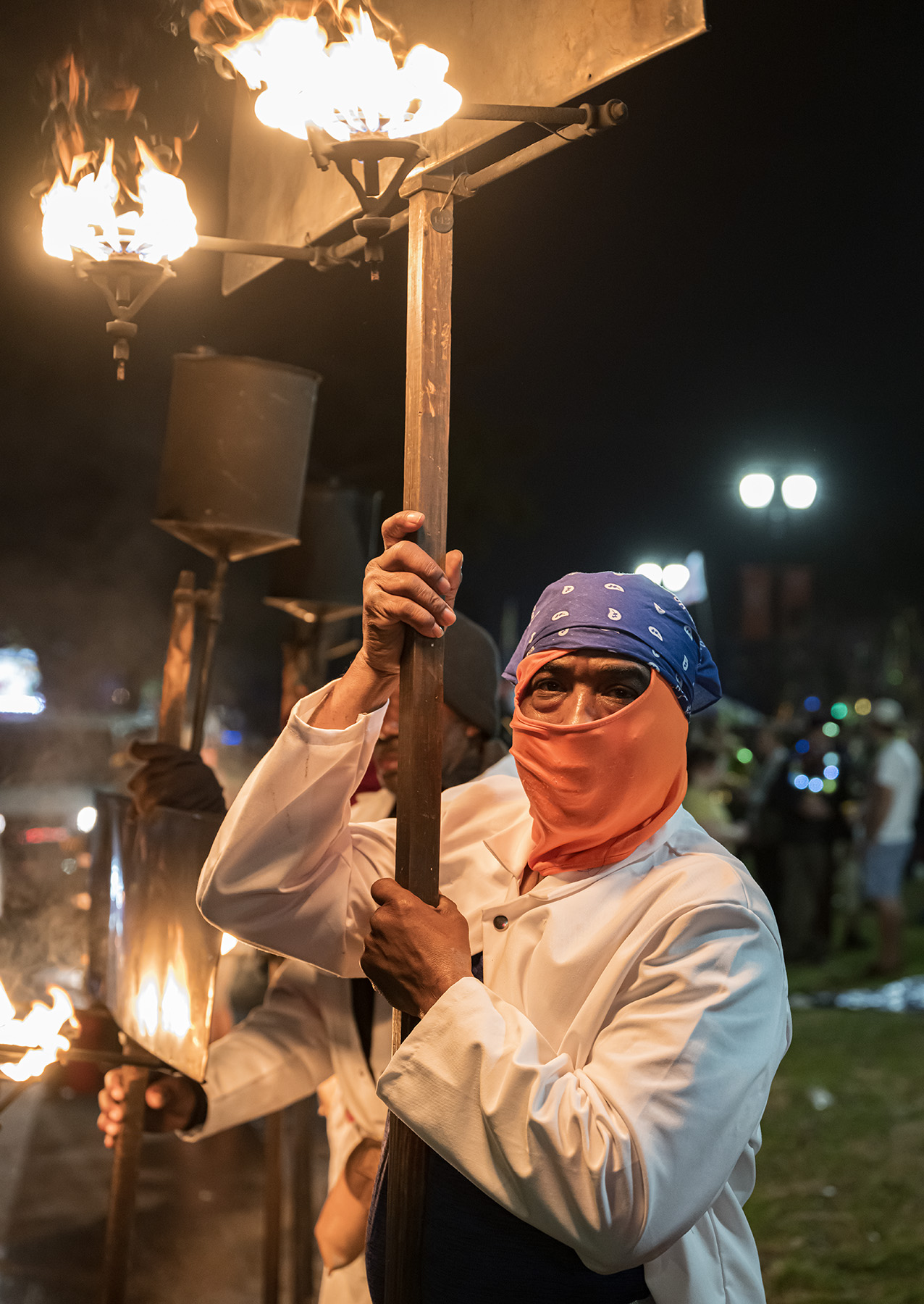 Person wearing a blue headscarf and orange face covering holding a torch with flames, standing near fire-lit structures at night.