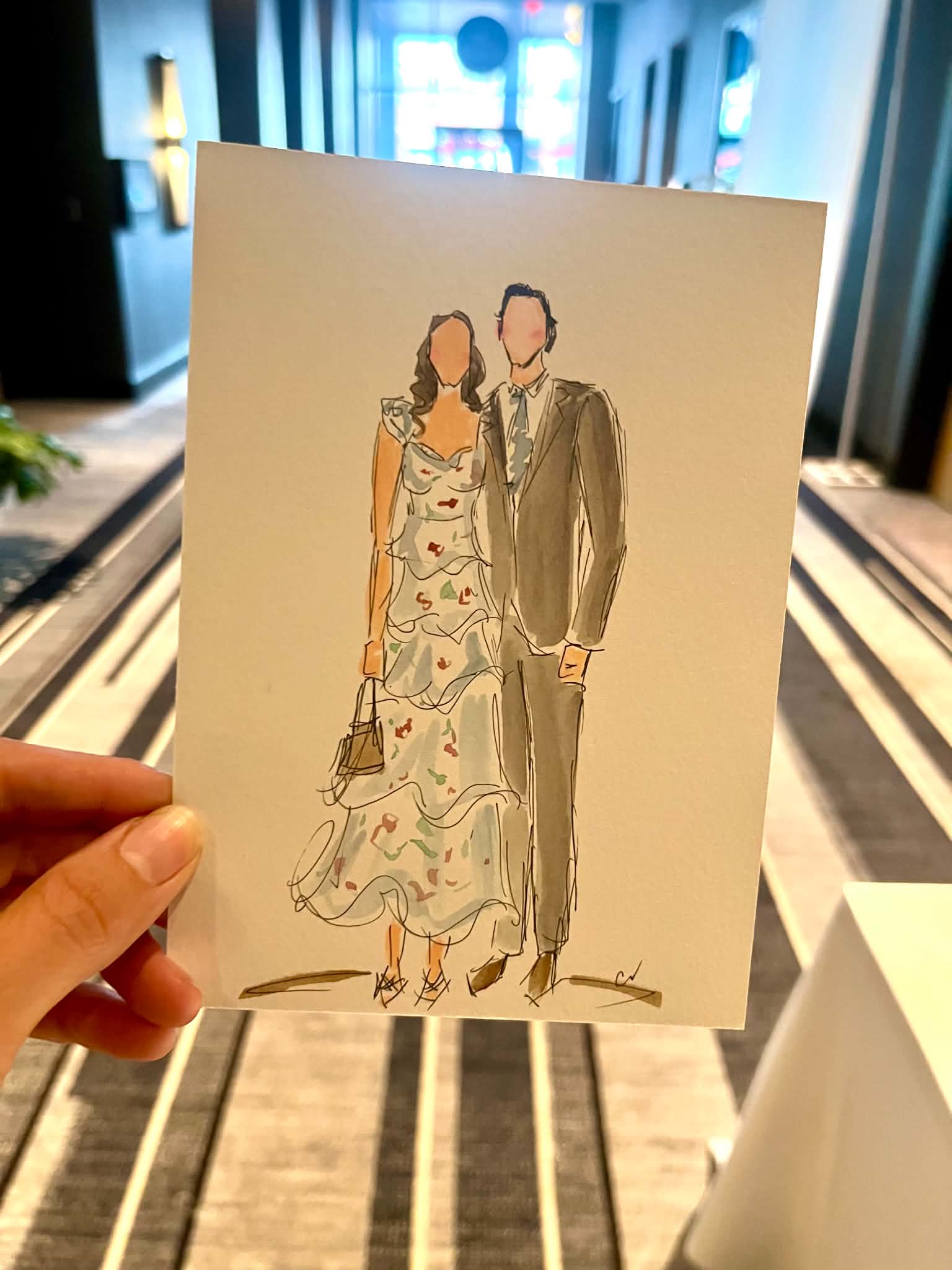 Hand holding a drawing of a woman in a floral dress and a man in a suit walking together, indoors with blue lighting.