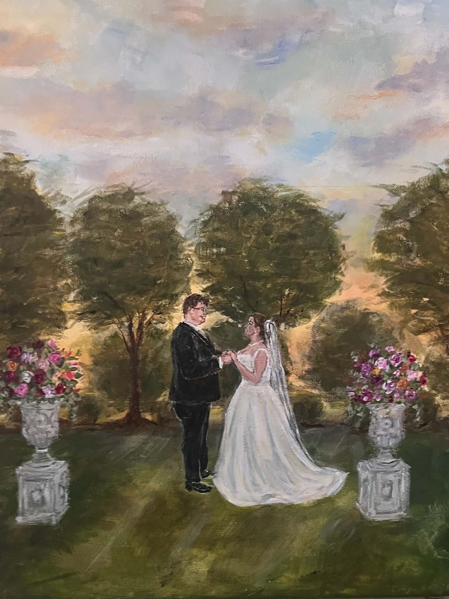 A bride and groom stand outdoors under trees, holding hands, with pink flowers in stone planters, during sunset.