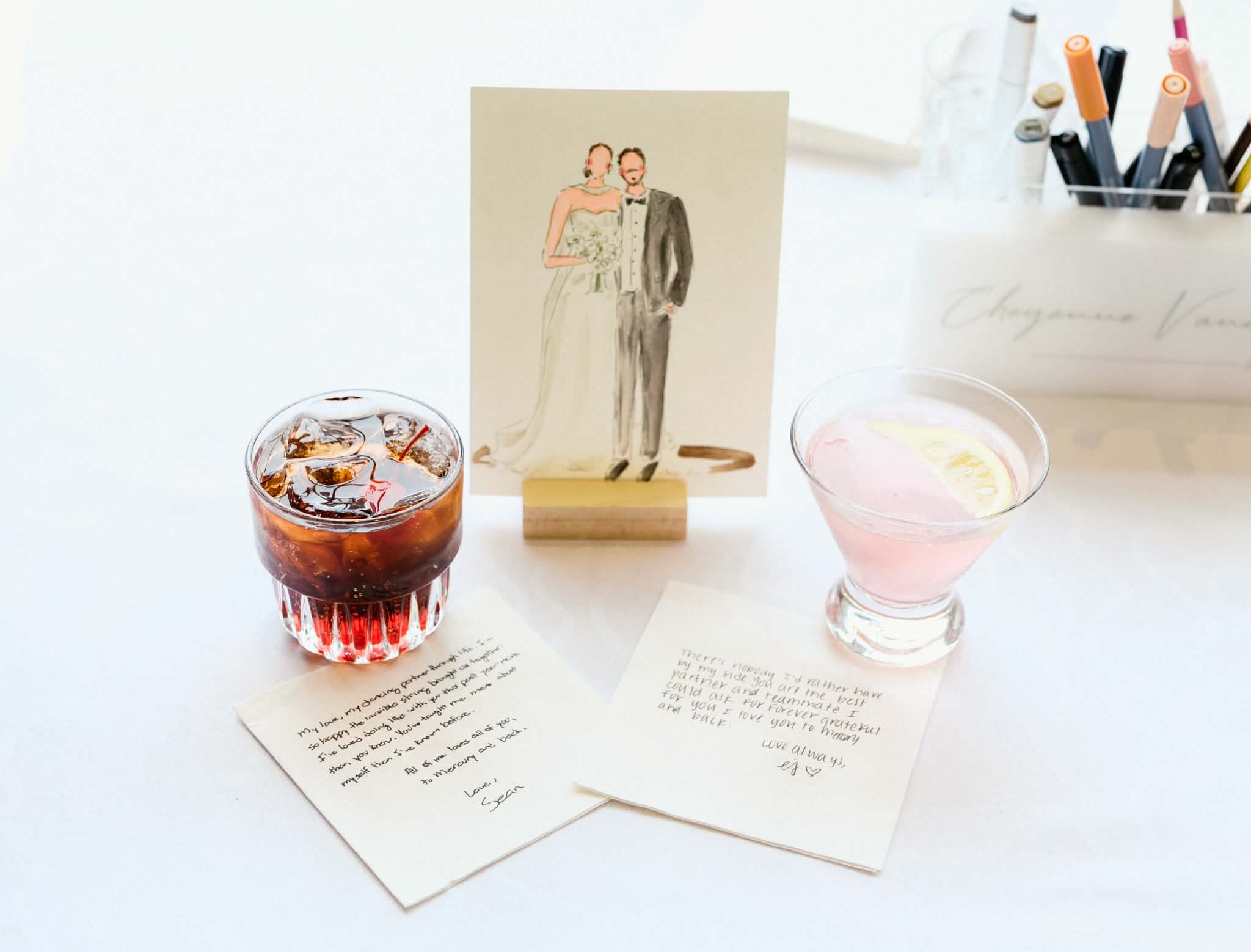 Wedding card with drinks and handwritten notes on a white table, with pens in a container in the background.