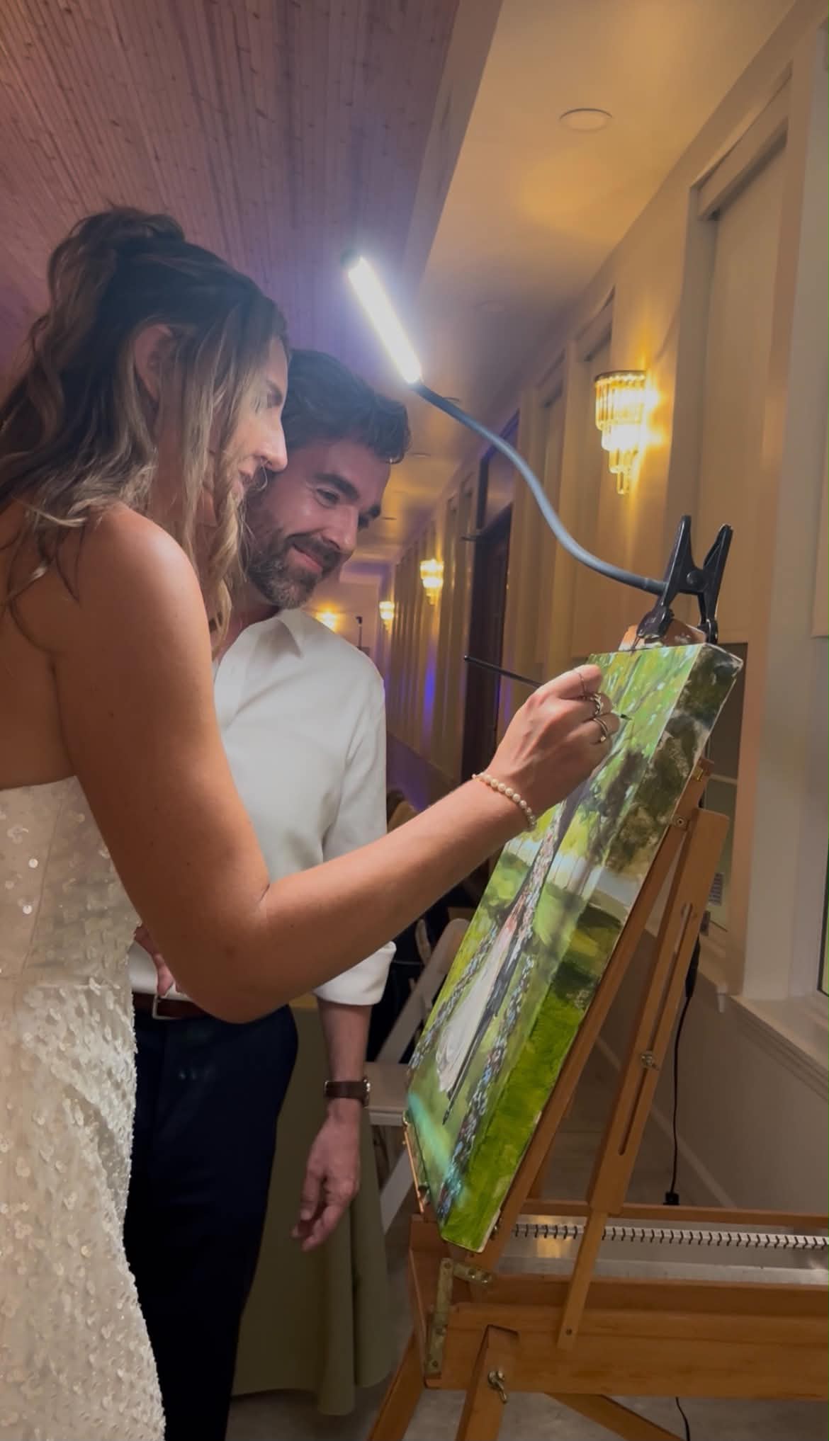 Two people, a woman and a man, stand near an easel with a painting, in a warmly lit room.