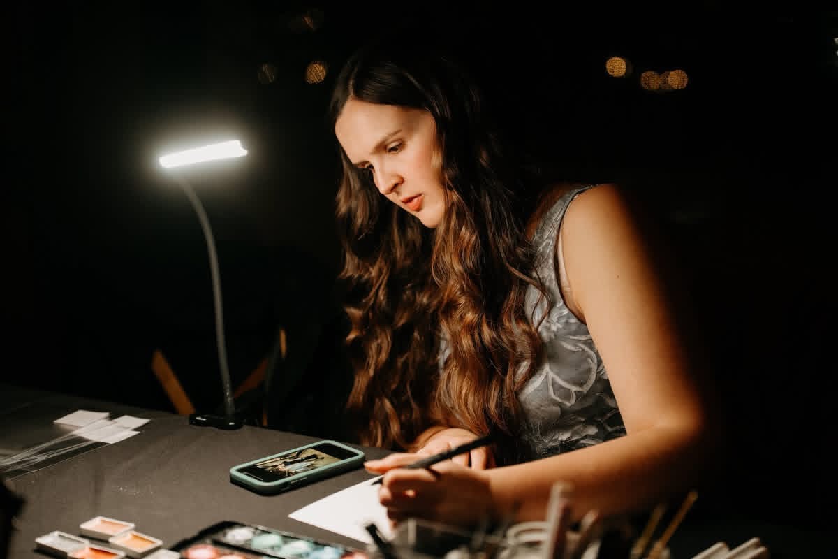 Woman with long wavy hair writing on paper at desk with phone and lamp, dark background, focused expression.