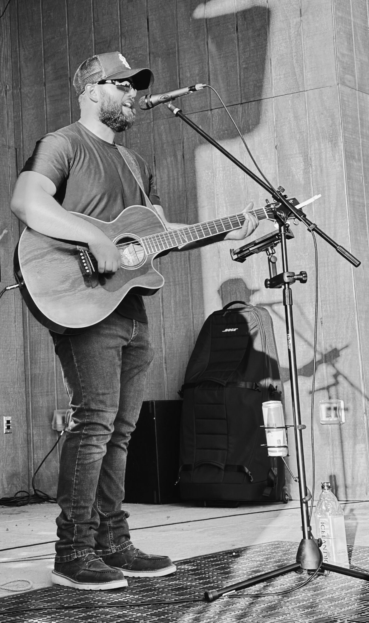 Man with a beard, wearing a cap, playing an acoustic guitar and singing into a microphone on stage.