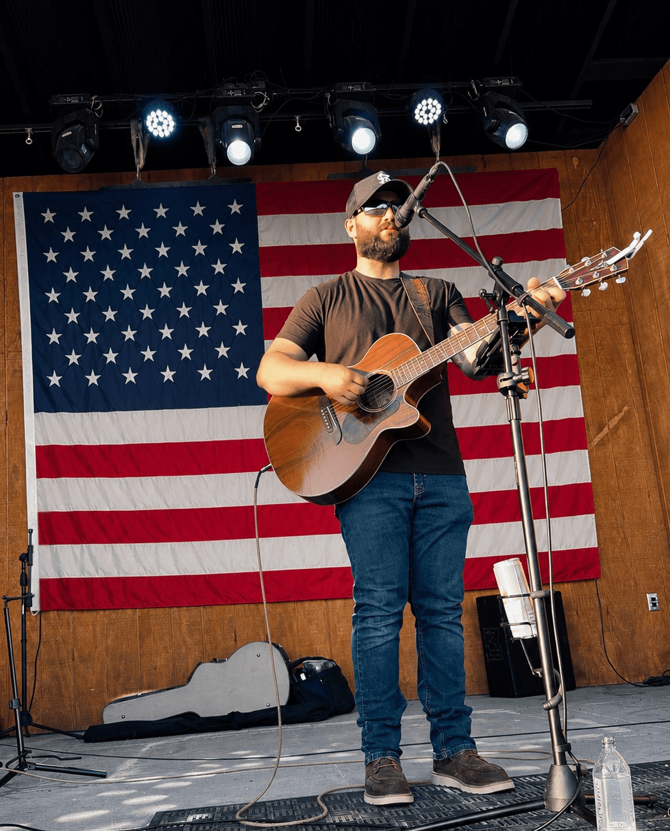 Man playing guitar on stage with American flag backdrop, stage lights above, and microphone in front, holding a guitar and wearing sunglasses.