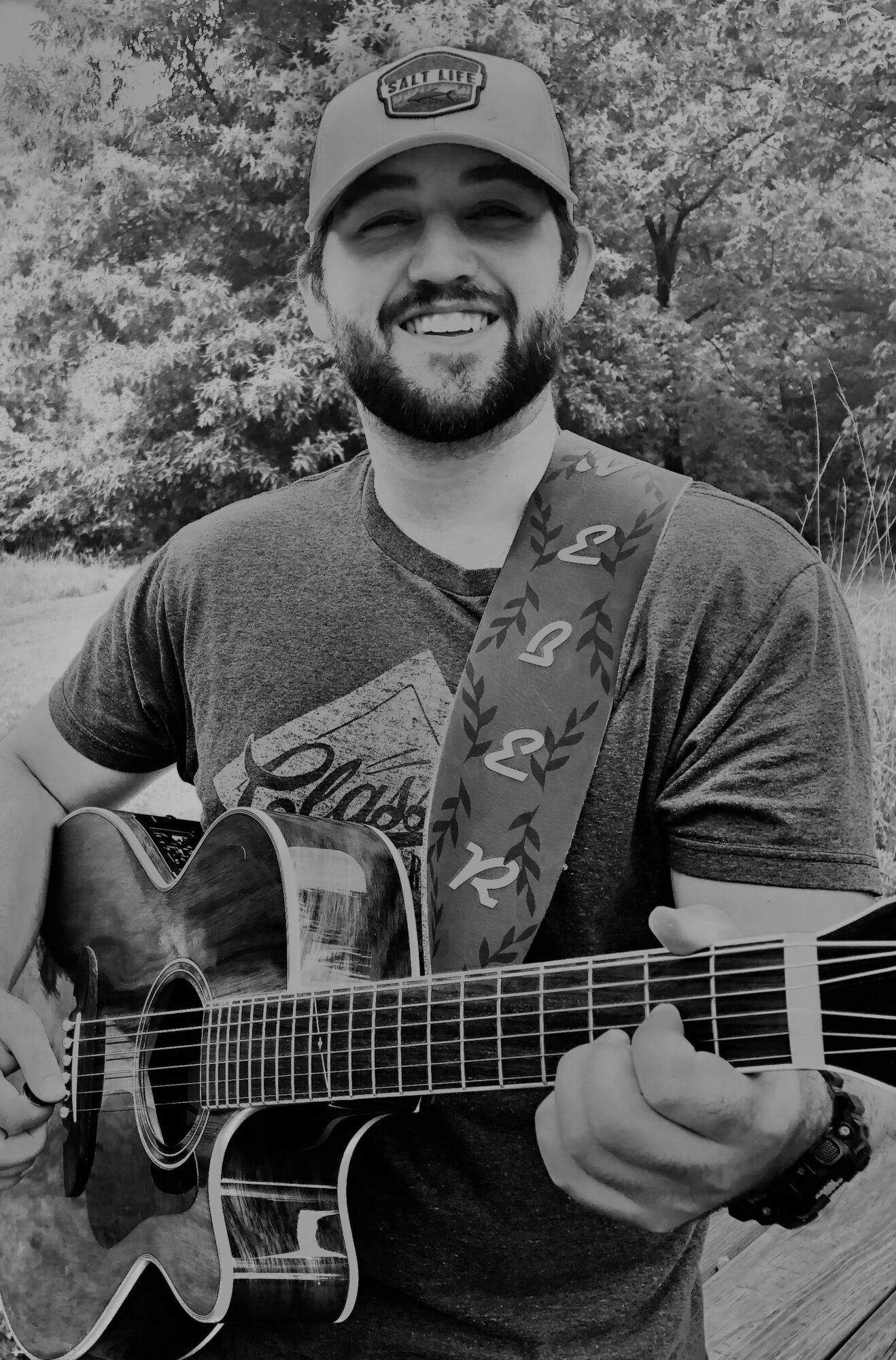 Man smiling, wearing a cap, playing an acoustic guitar outdoors with trees in background.