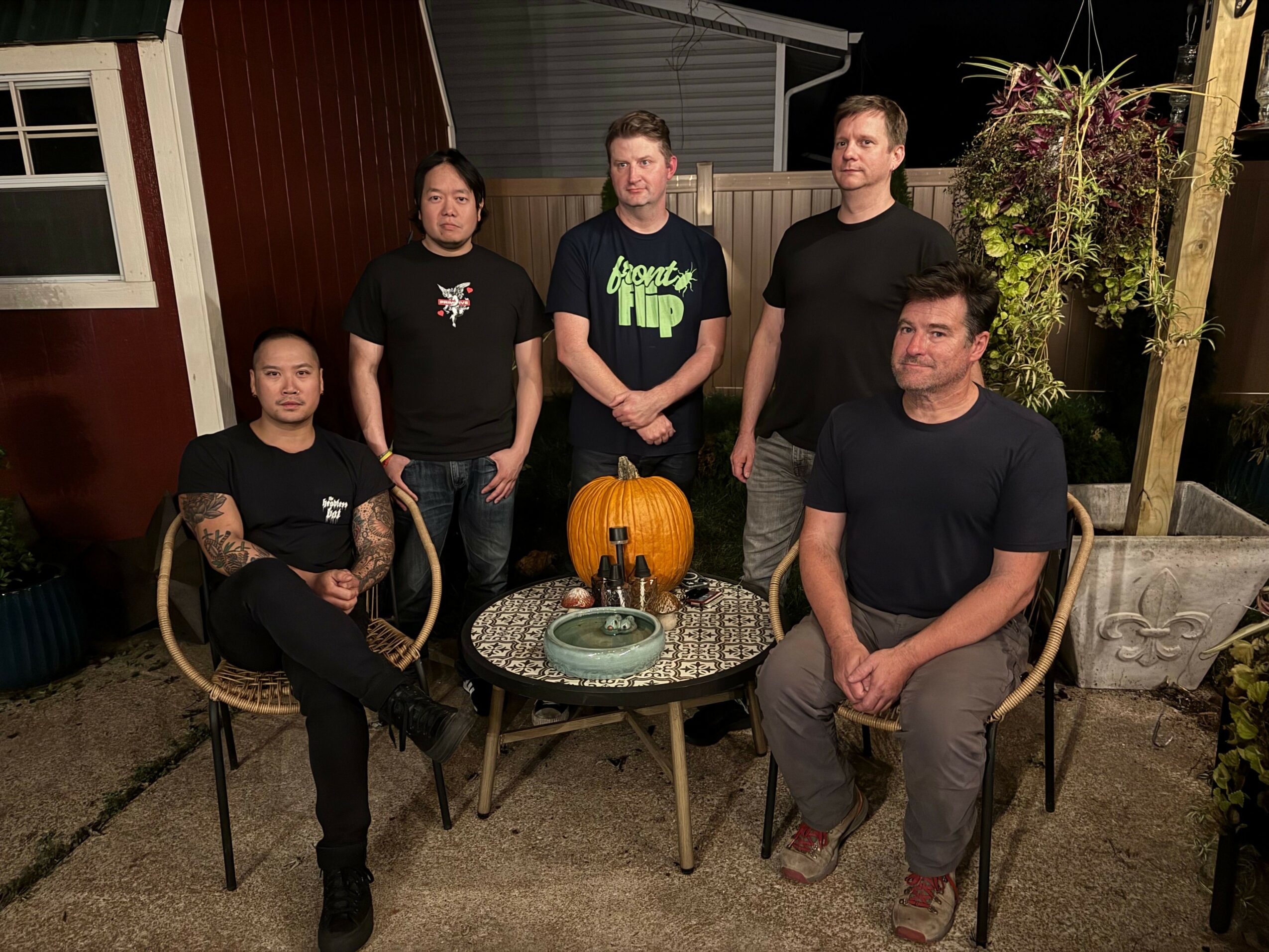 Four people sitting around a table on a patio, with a pumpkin decoration, plants, and a building in the background.