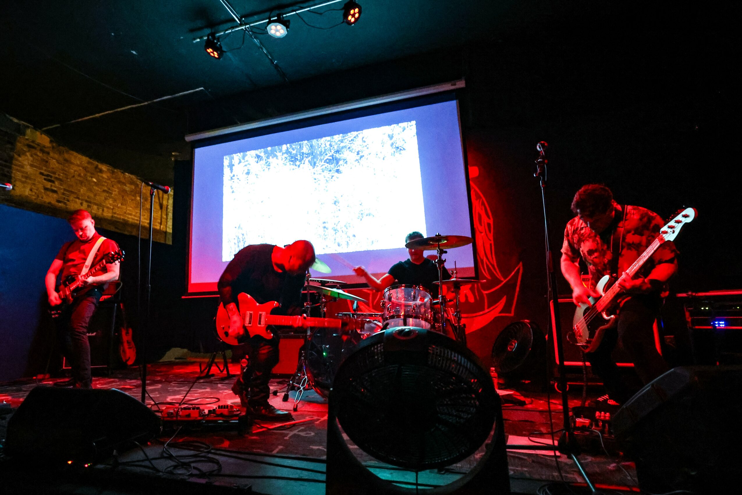 Band performing live on stage with instruments, screen in background, dim lighting, and red accents.