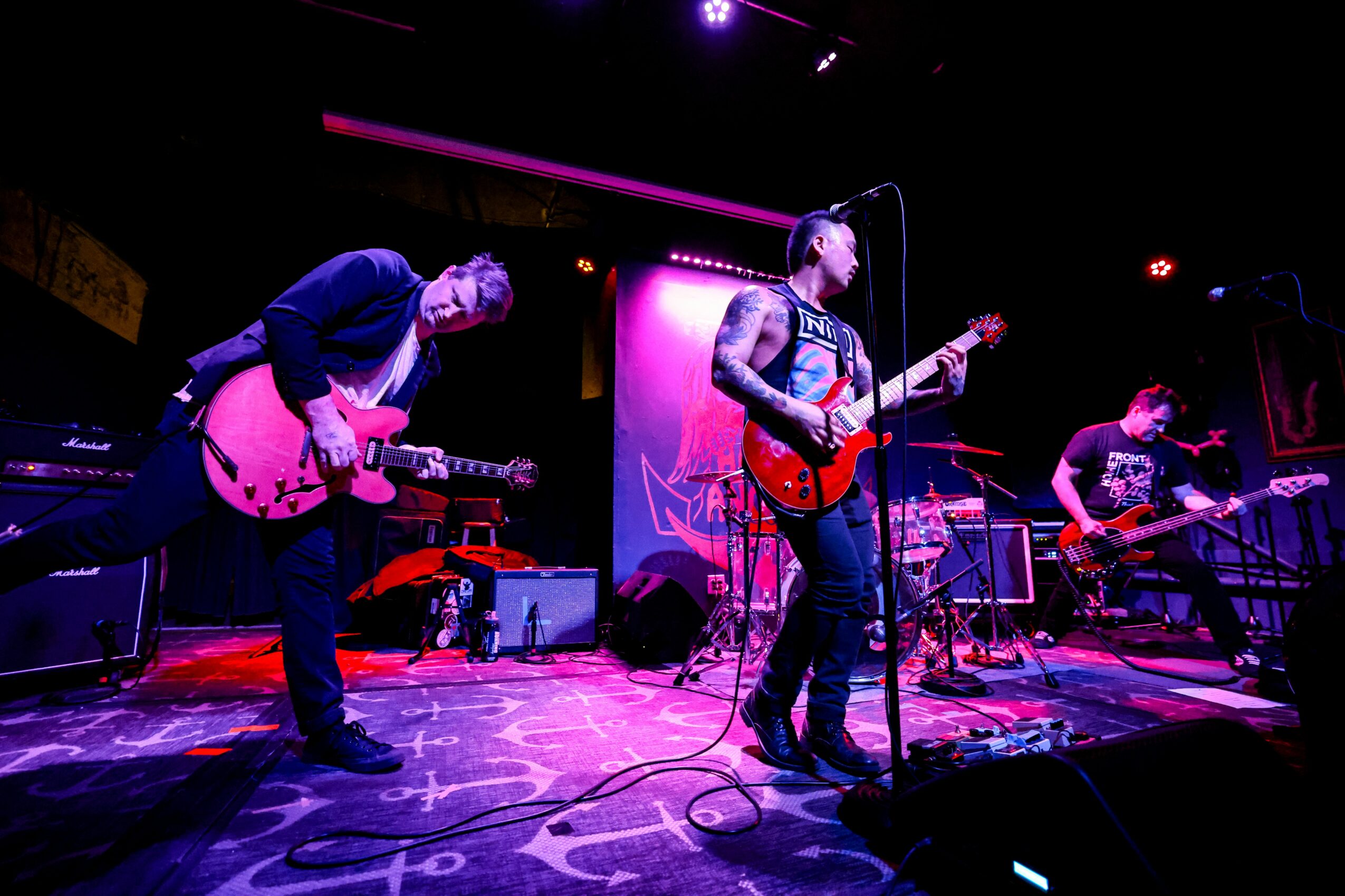Band performs on stage with three guitarists, colorful lighting, and musical equipment visible, audience not shown.