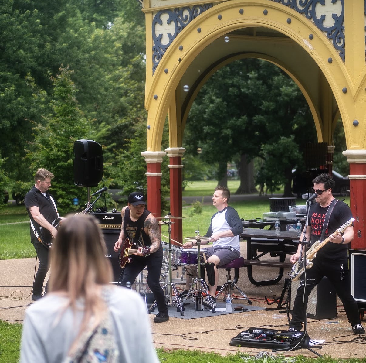 Band performing outdoors under a decorative yellow and blue arch with trees in the background.