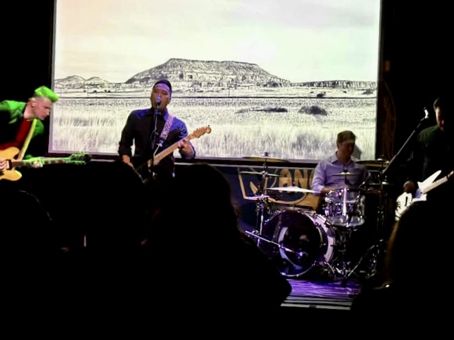 Band performing on stage with a black-and-white landscape background, including a mountain and open land, behind them.