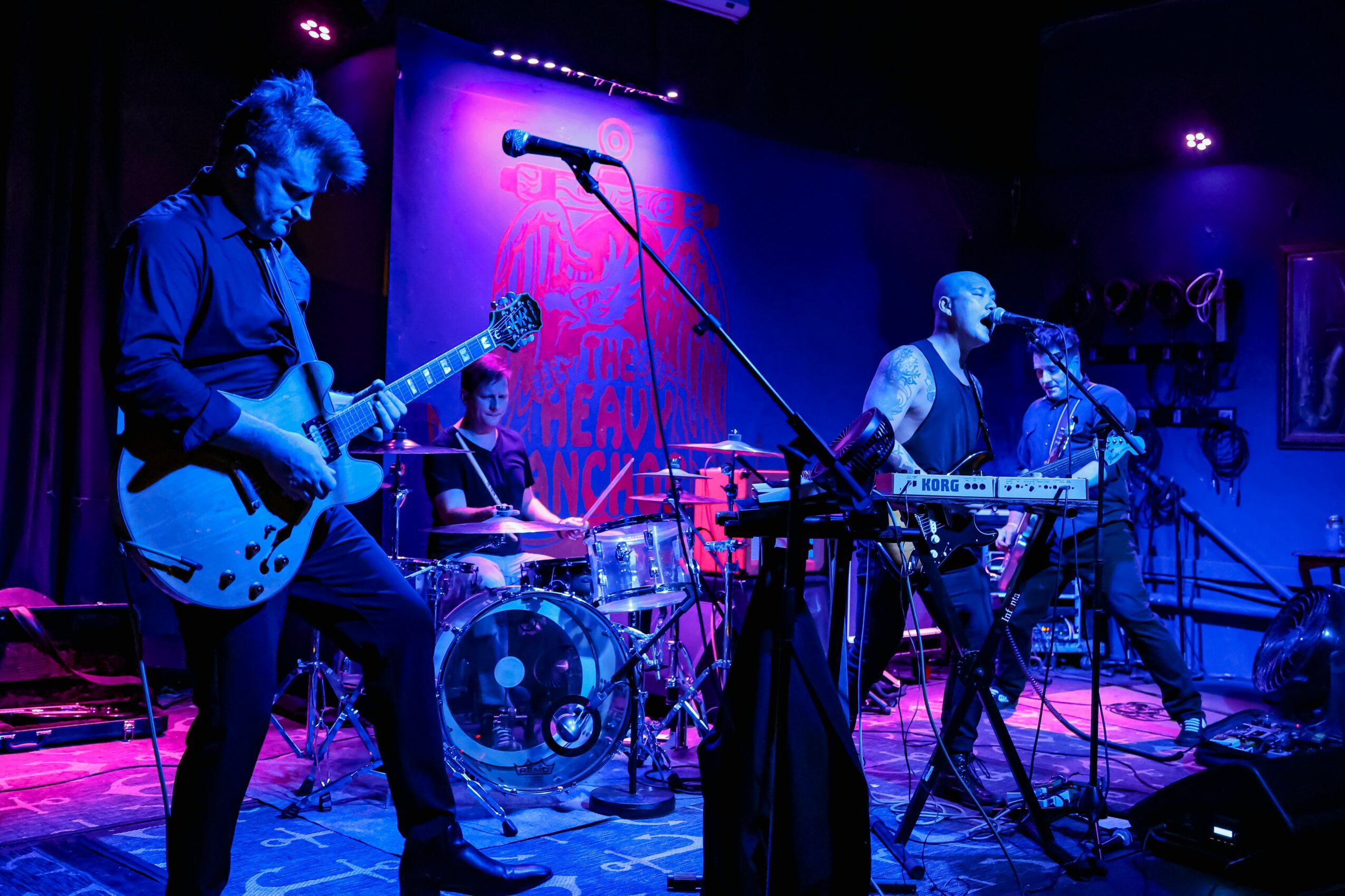 Band performing on stage with guitar, drums, keyboard, and microphone under purple and blue lighting.