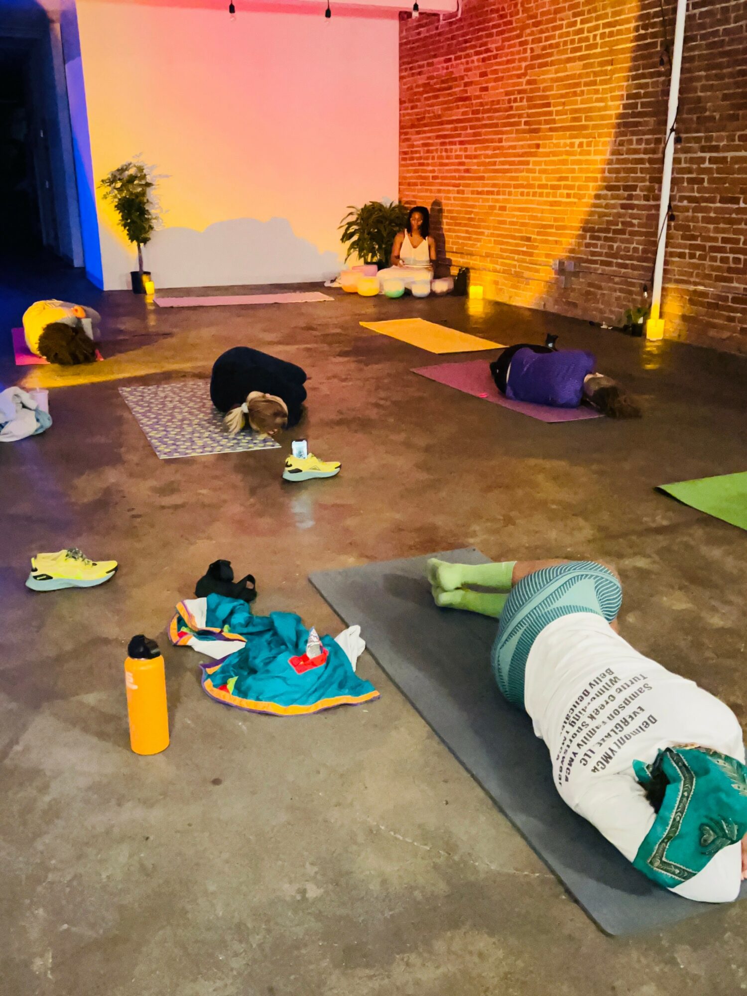 People practicing yoga on mats in a spacious room with brick and painted walls, some with water bottles nearby.