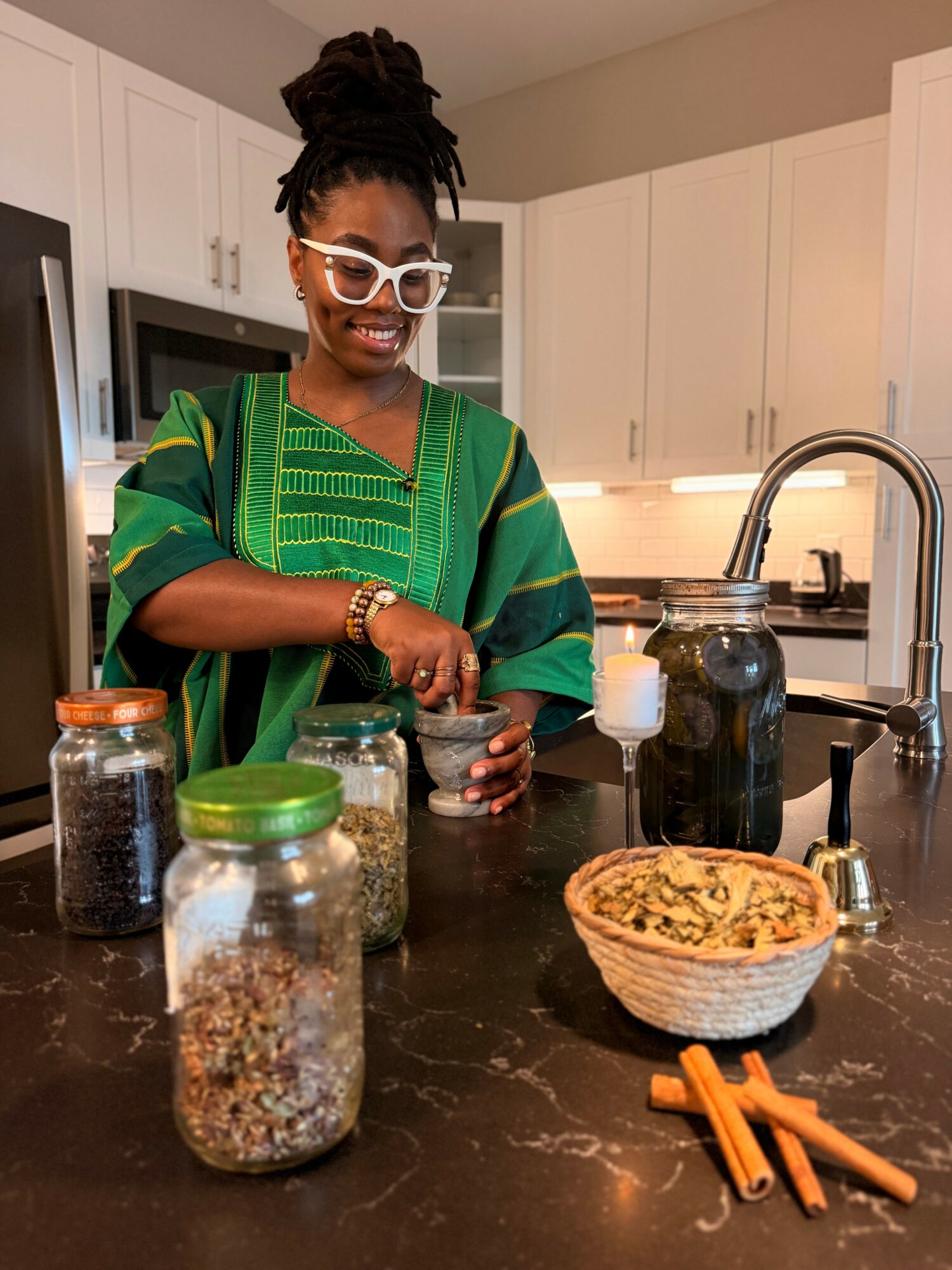 Woman with glasses and dreadlocks preparing food in a kitchen, jars and a bowl on the counter.