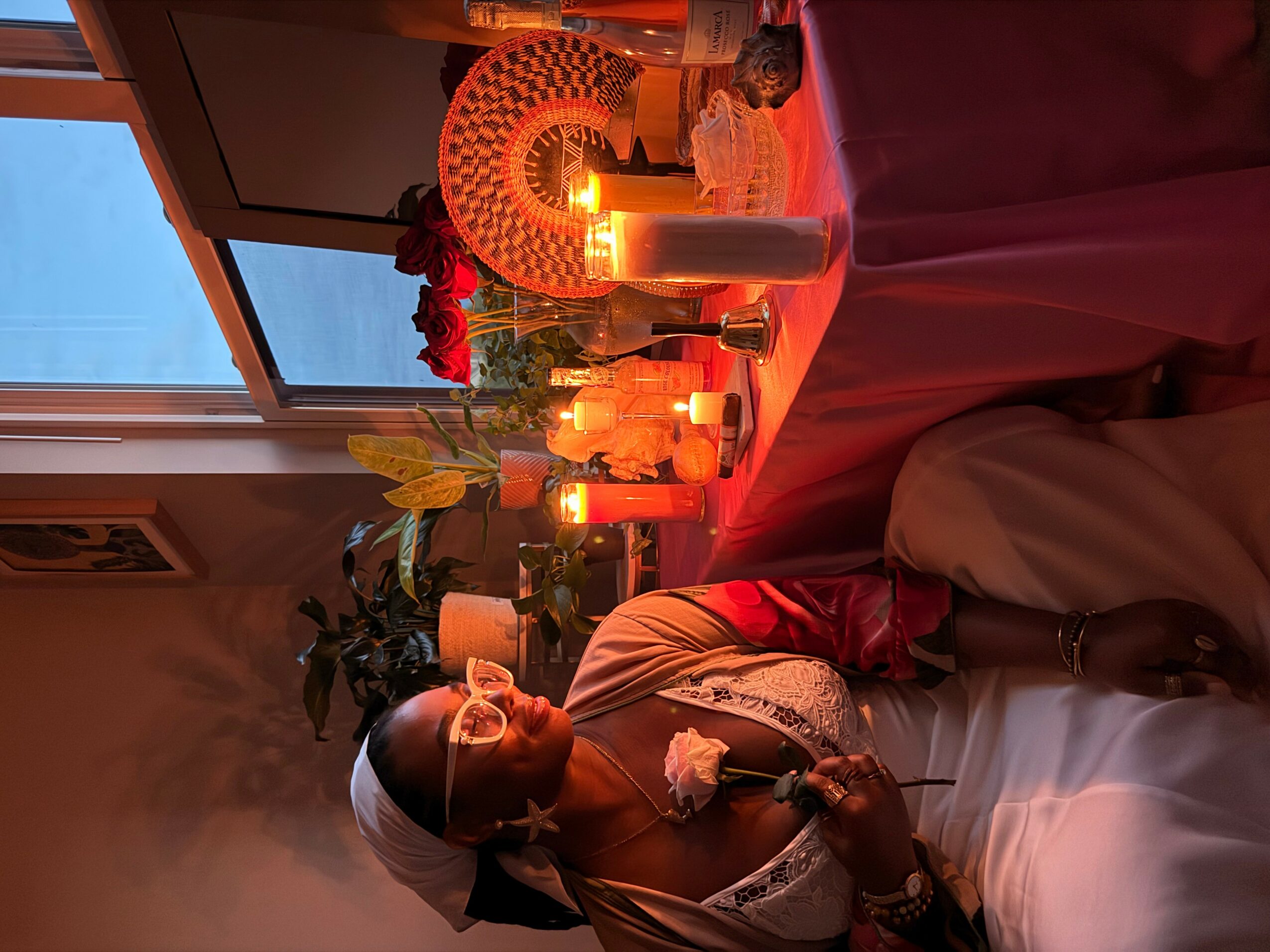 Person sitting at a table with lit candles, flowers, and decorative items, near a window with a view outside.
