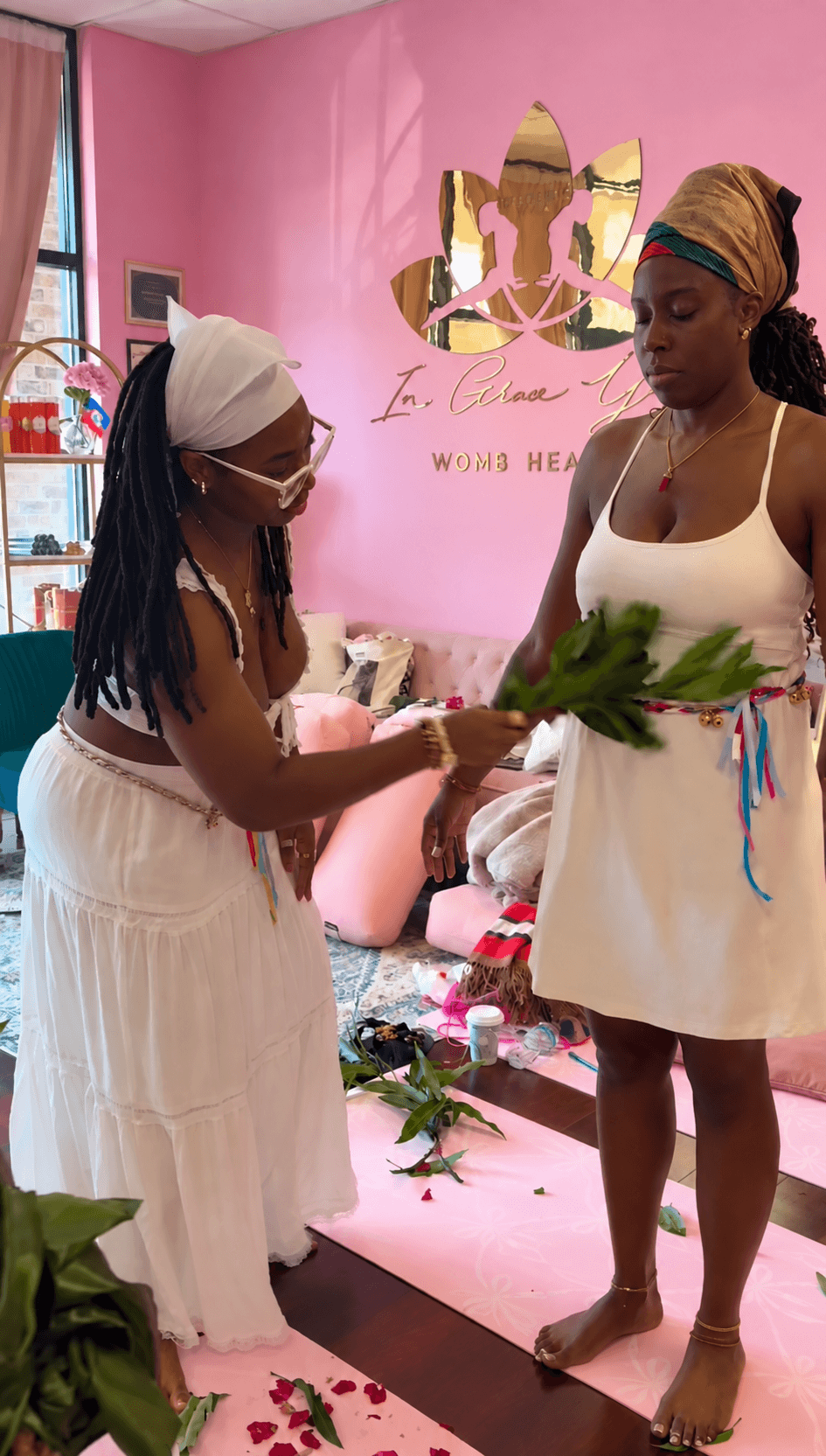 Two women in a pink room exchanging flowers, one wearing a white dress and headwrap, the other in a white dress with a headscarf.