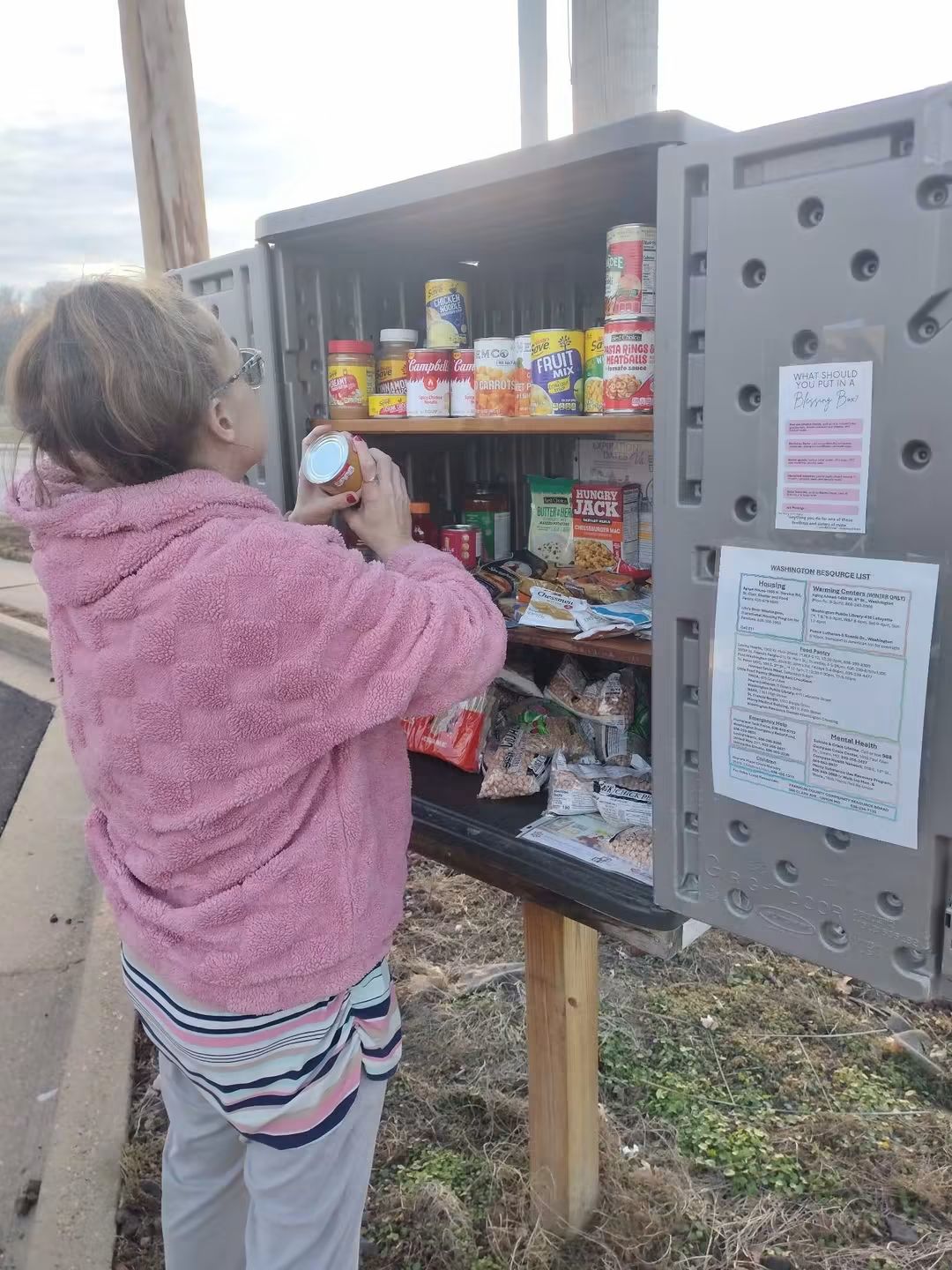 Person in pink jacket and striped pants looking at a shelf of canned and packaged food in an outdoor storage box.