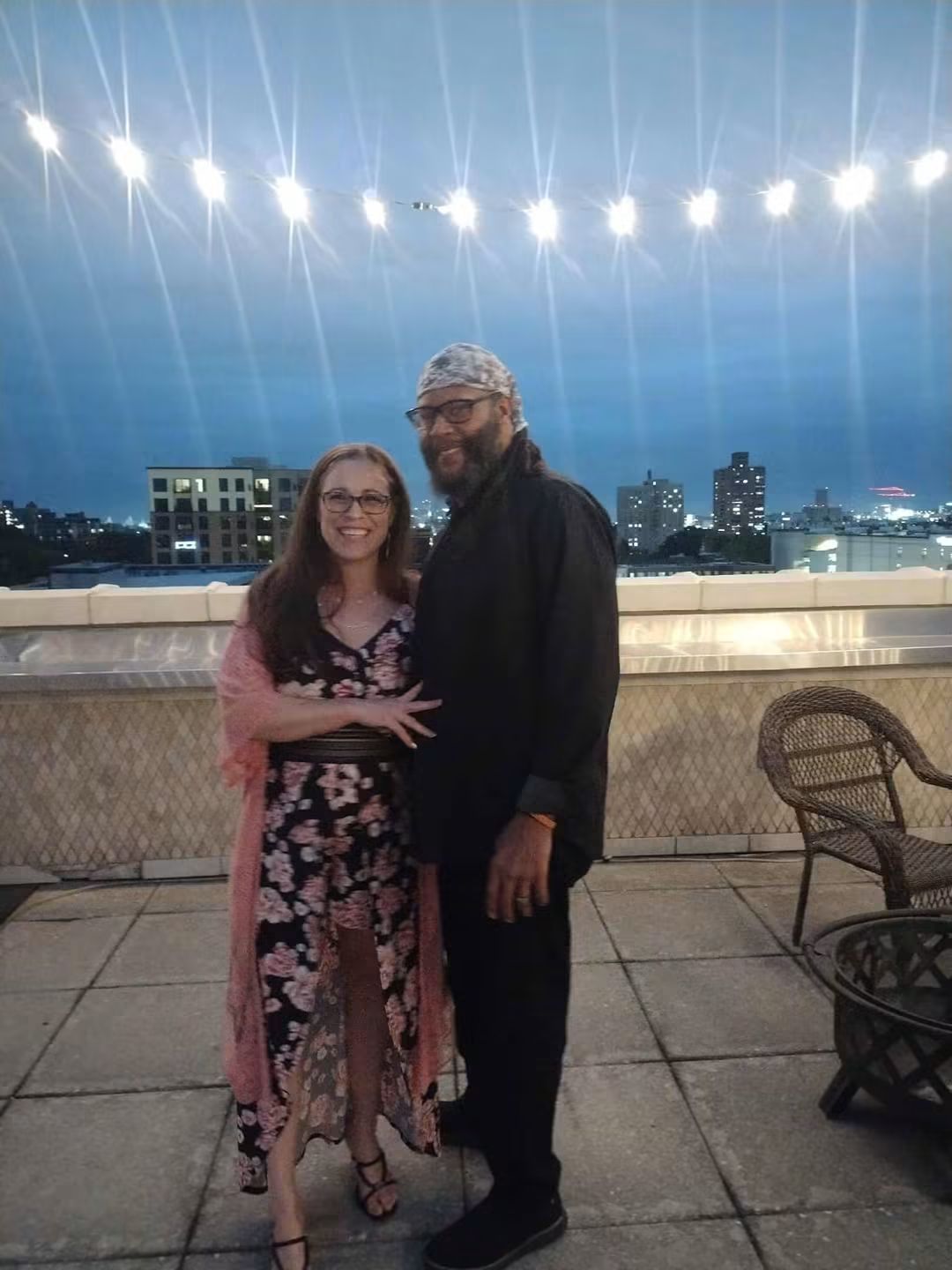 A woman and a man stand together on a rooftop at night, with city buildings and string lights in the background.
