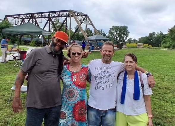 Four people standing outdoors on grass, smiling, with tents and trees in the background.