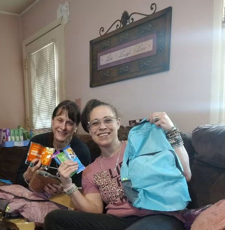Two women sitting on a couch, smiling, holding snacks and a blue backpack in a cozy room with a window and wall decor.