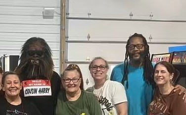Group of five people standing indoors in front of a garage door, smiling, with bookshelves visible on the right.