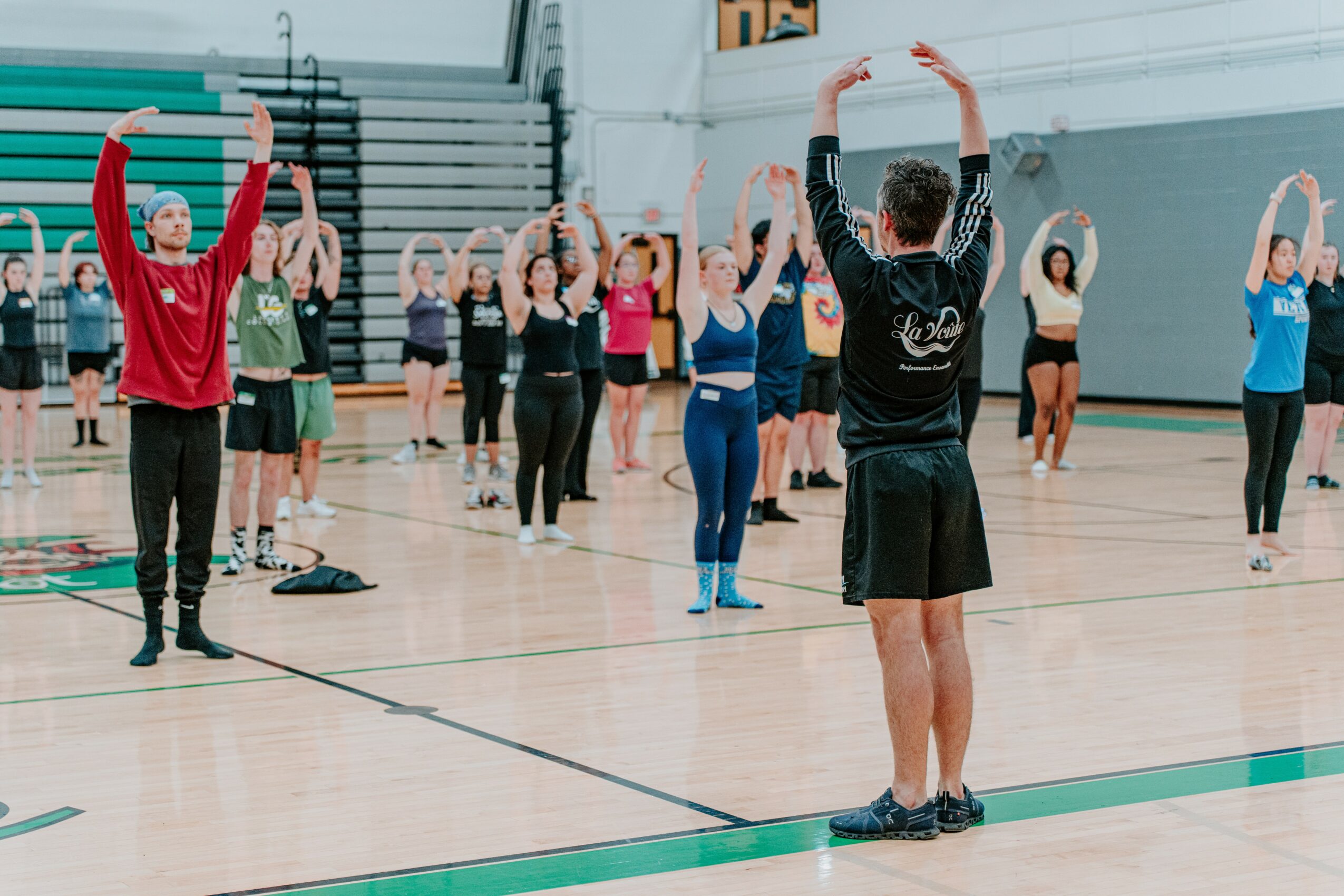 Group of people participating in a fitness class in a gymnasium, following instructor's lead with arms raised.