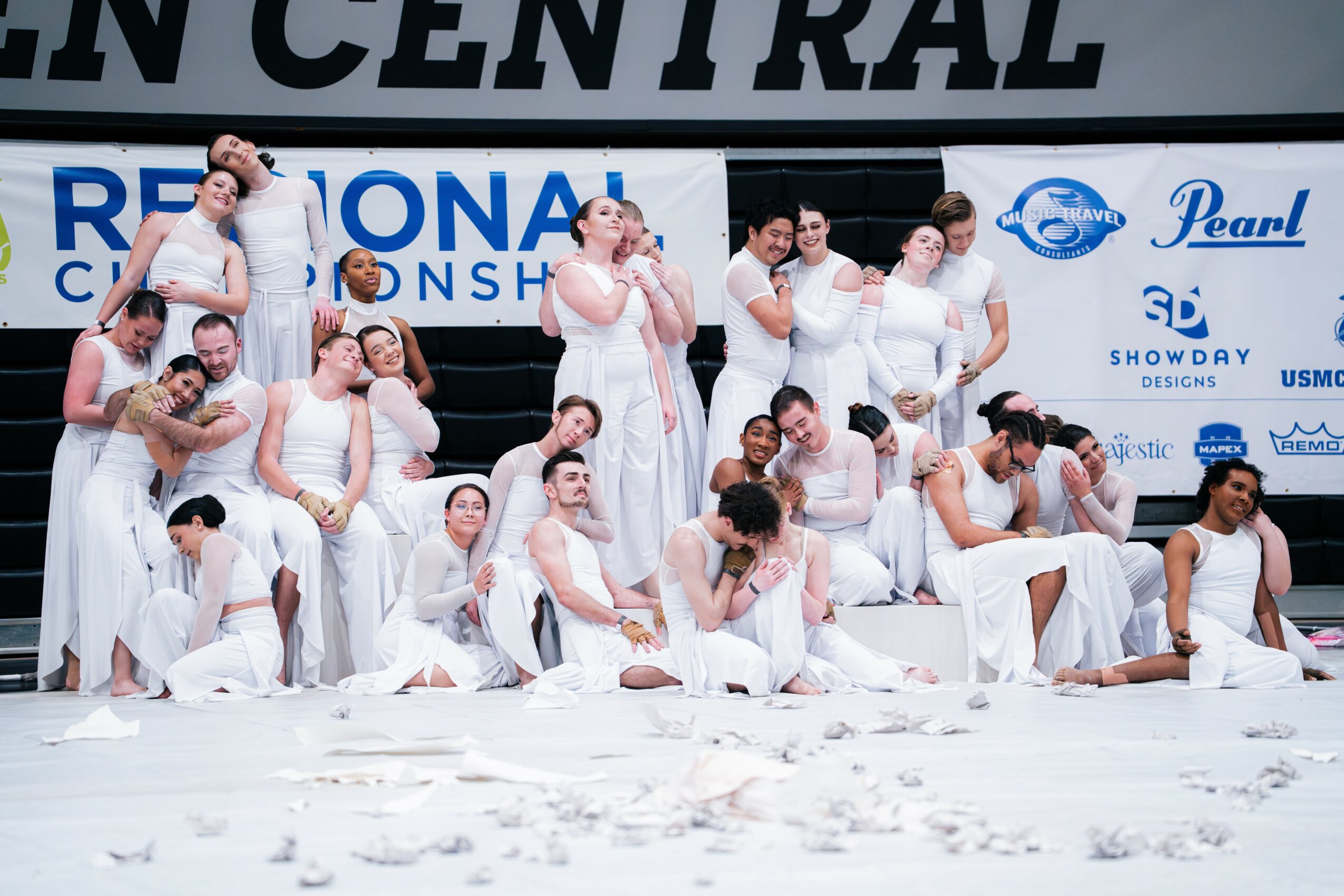 Group of people dressed in white costumes on stage, some sitting, some standing, with a backdrop displaying text and logos.