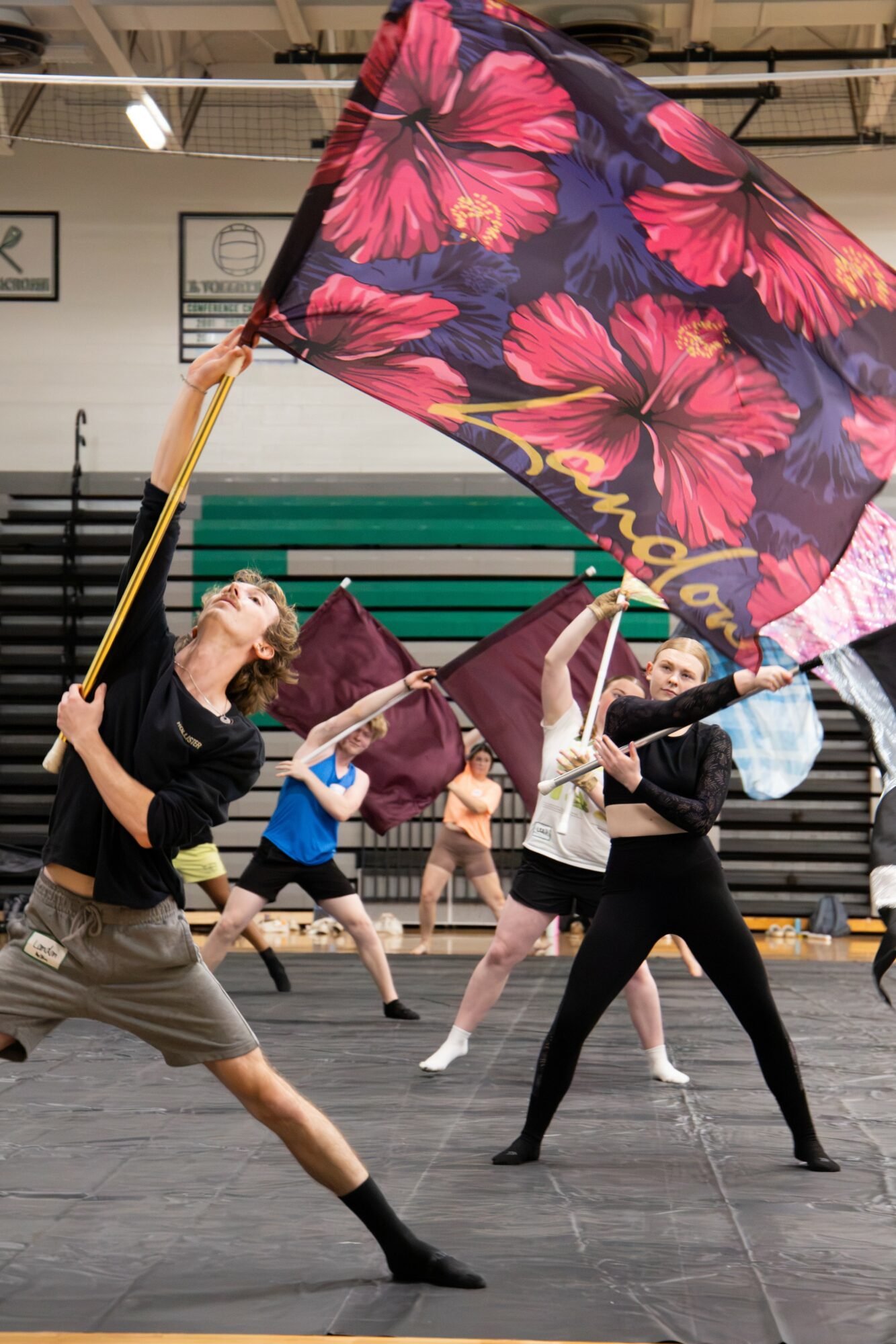 Person holding a large floral flag in a gymnasium with other performers in the background.