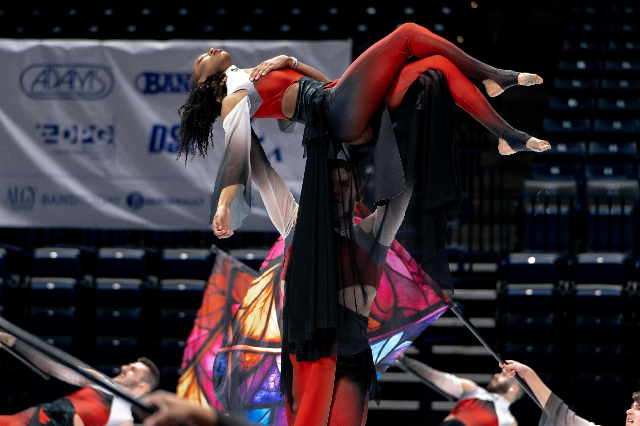 Performer lifts a person during a dance on stage with a colorful backdrop and audience seating.