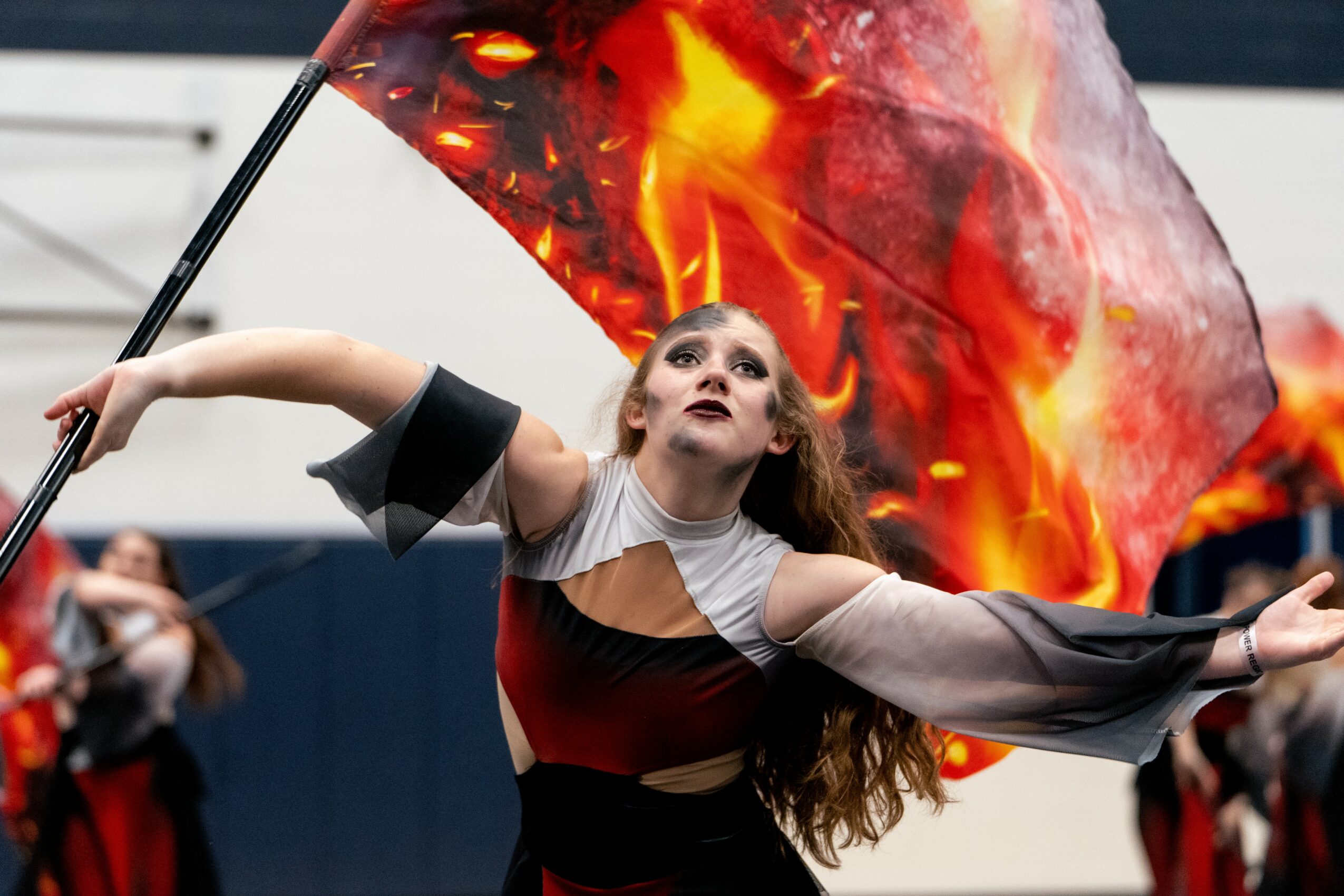 A performer with a flag featuring flames, arms outstretched, looking upward during a performance, with blurred audience in background.