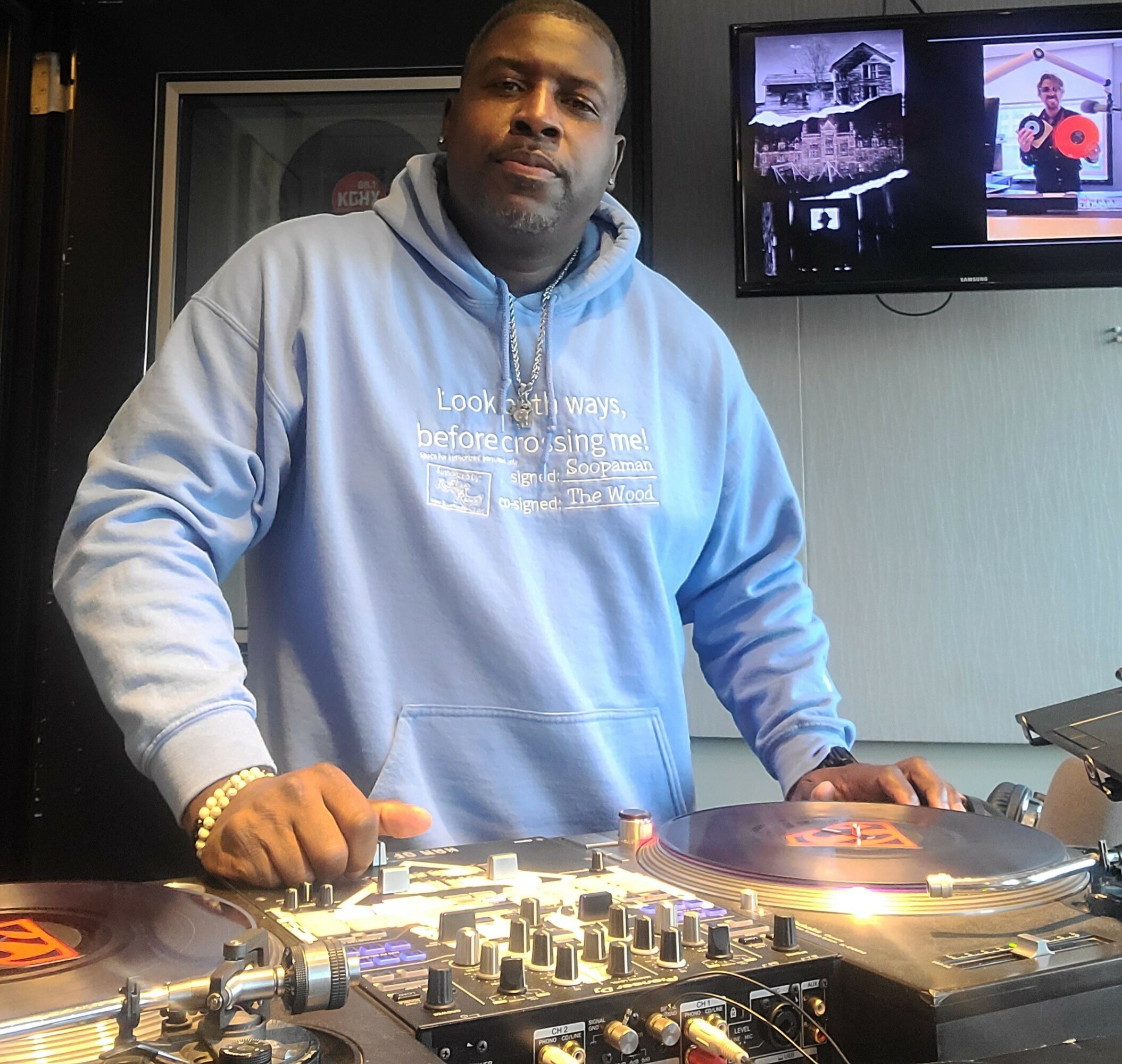 Man in a light blue hoodie standing behind DJ equipment, with a TV screen in the background displaying images.