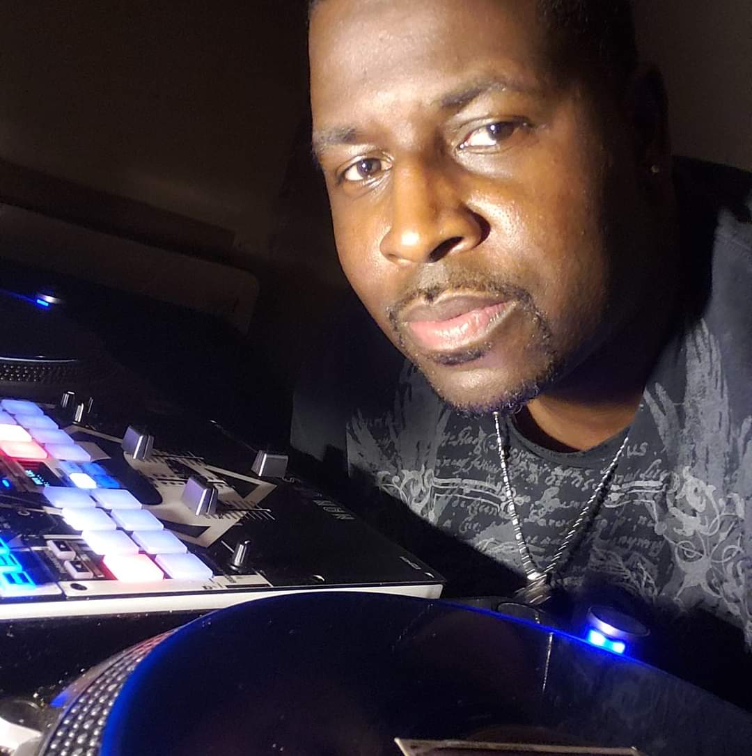 Person with dark skin and short hair looking at the camera, with DJ equipment in the foreground.