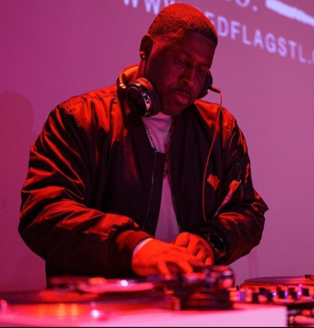 DJ wearing headphones and a black jacket, operating turntables in a dimly lit setting with pink and purple lighting.