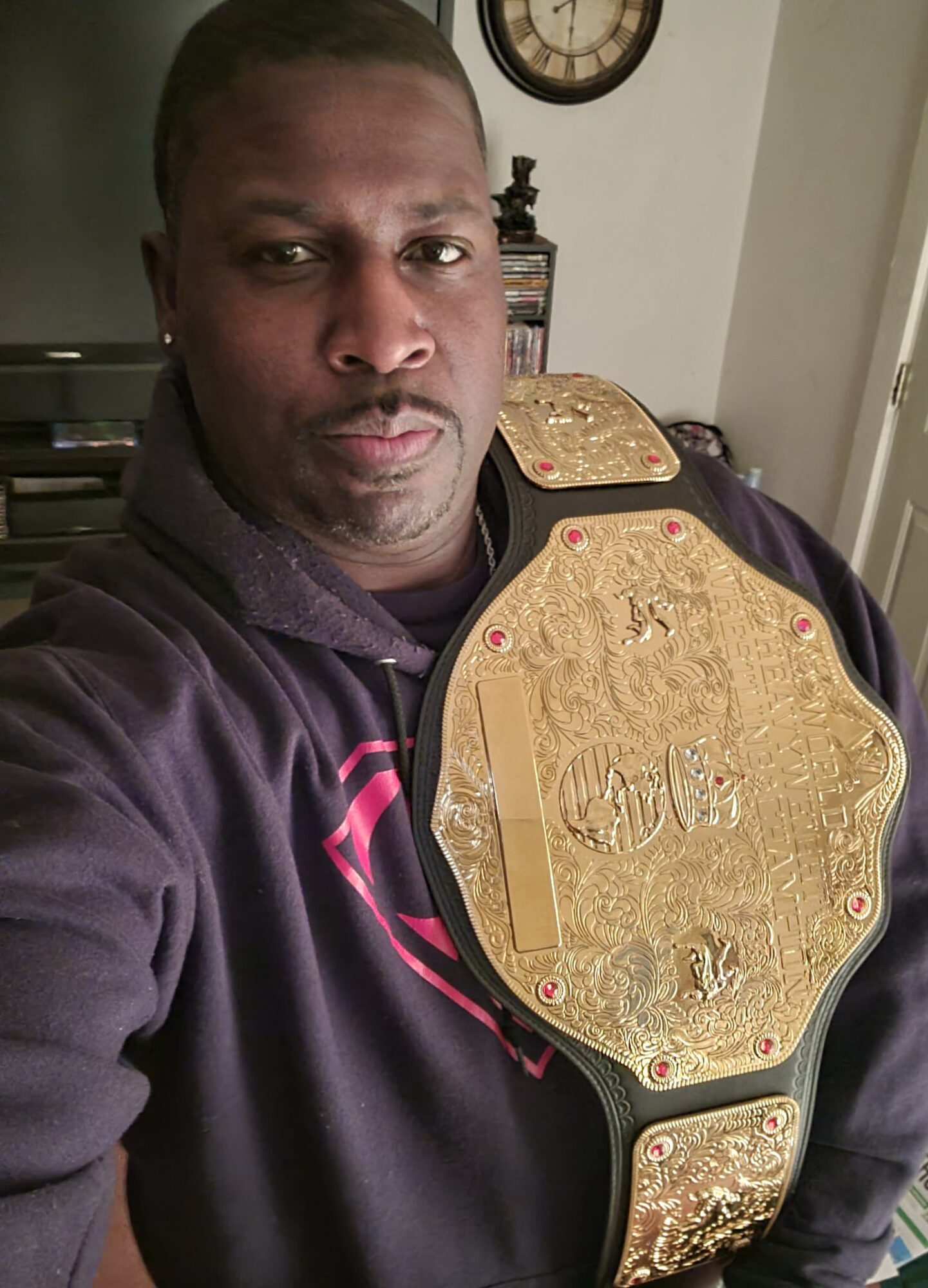 Person taking a selfie wearing a large gold championship belt over a dark hoodie, standing indoors with a clock and furniture in background.