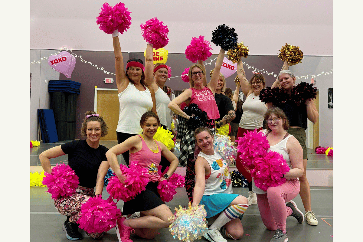 Group of women holding pink, black, and white pom-poms, celebrating in a decorated room with banners and balloons.