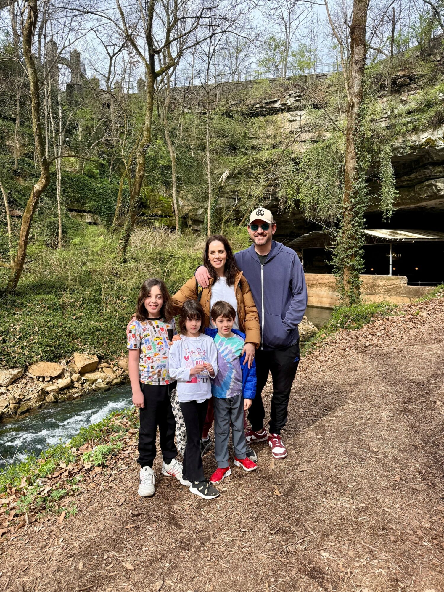 Group of five people standing on a dirt path near a stream with trees and a rock formation in the background.