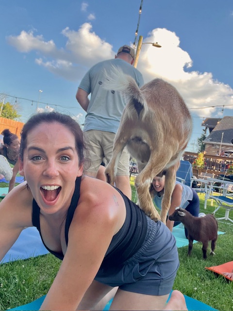 Woman smiling and leaning forward outdoors with a goat on her back, other goats and people in background, sunny day.