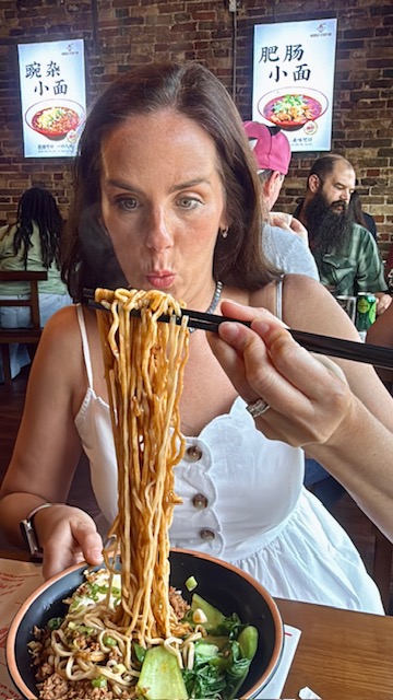 Woman eating noodles with chopsticks in a restaurant, holding a bowl of noodles, with posters on the wall behind her.