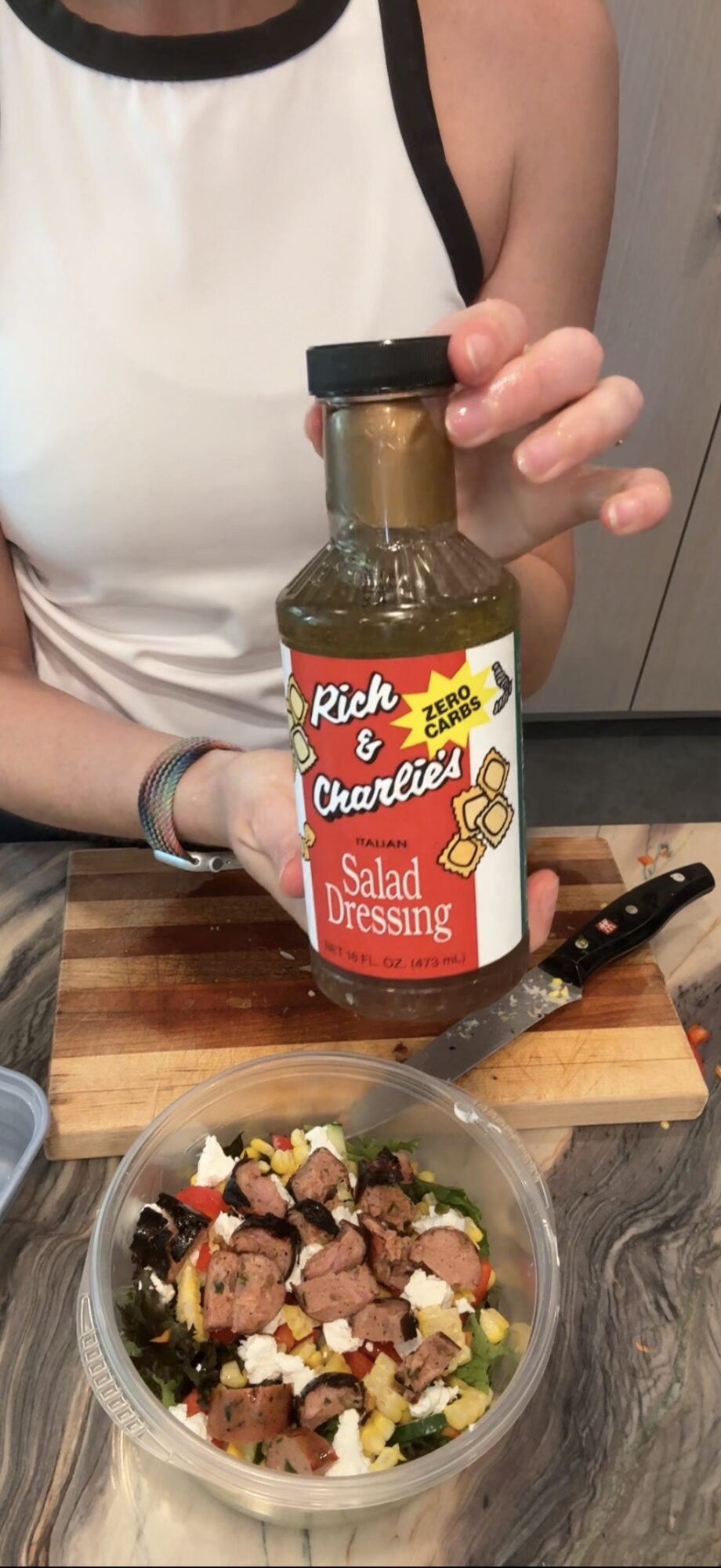 Person holding a bottle of salad dressing above a bowl of salad with various ingredients, on a kitchen counter