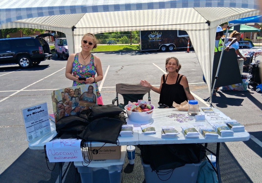 Two women stand behind a table under a canopy at an outdoor event, with various items displayed on the table.