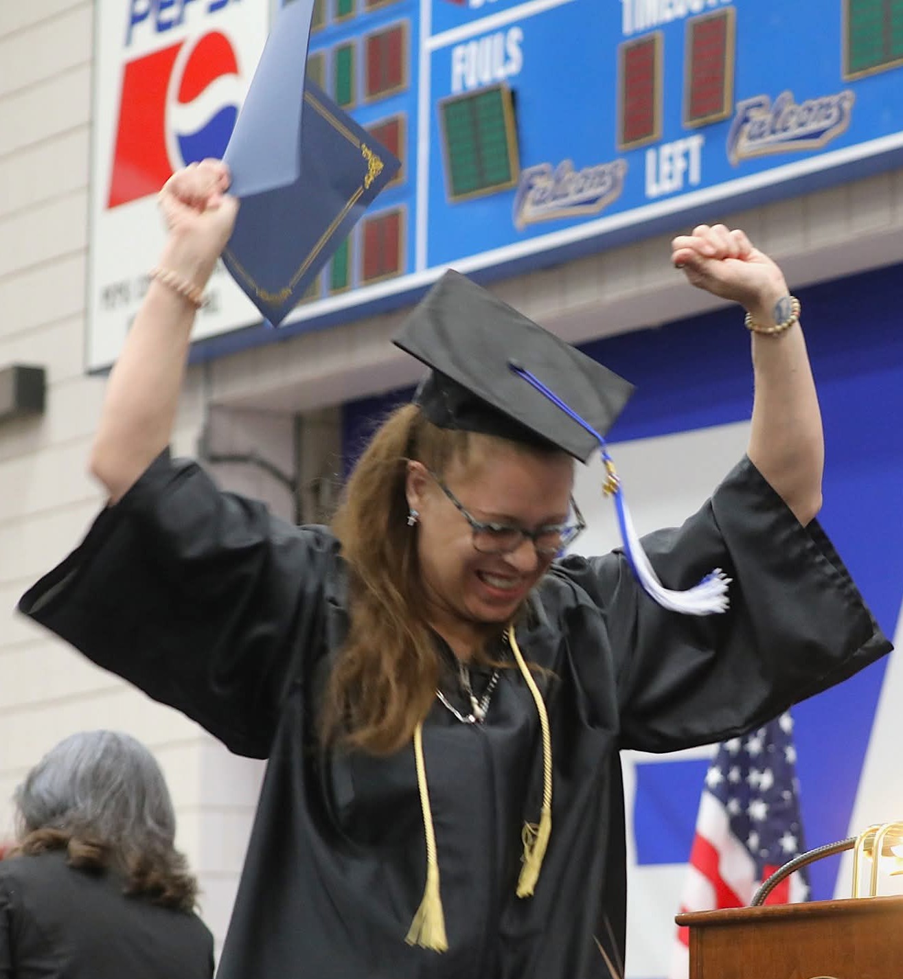 Woman in graduation gown and cap celebrating with arms raised, smiling, in front of a scoreboard and American flag.