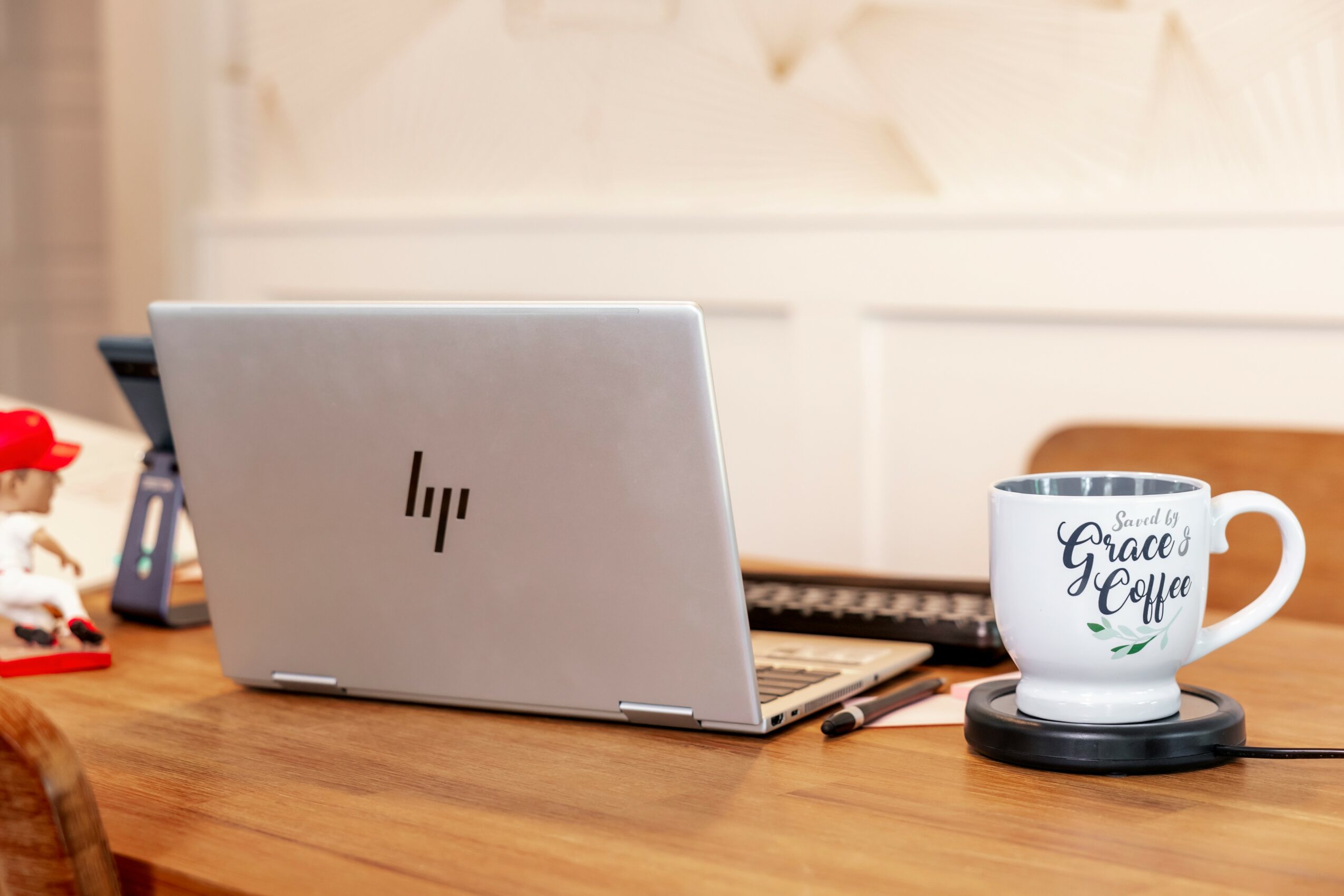 Laptop, coffee mug on a wooden table, blurred background, with a toy and a phone nearby.