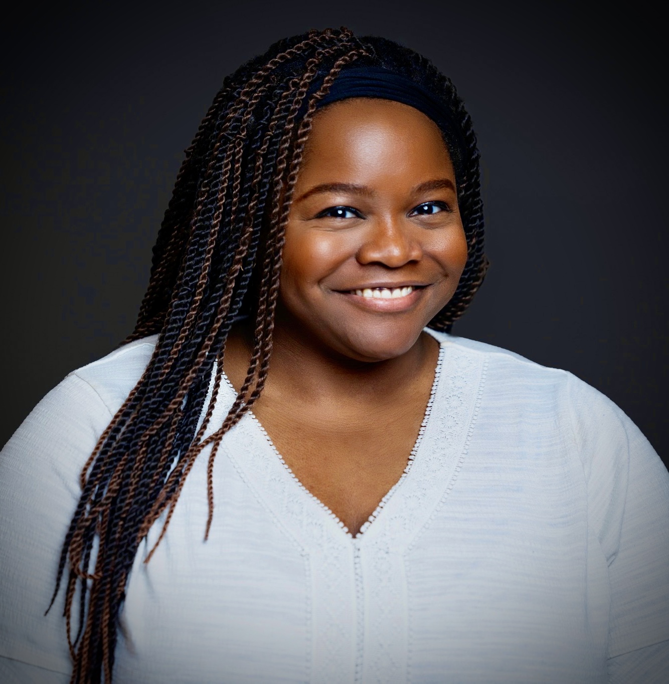 Smiling woman with braided hair wearing a white top against a dark background.