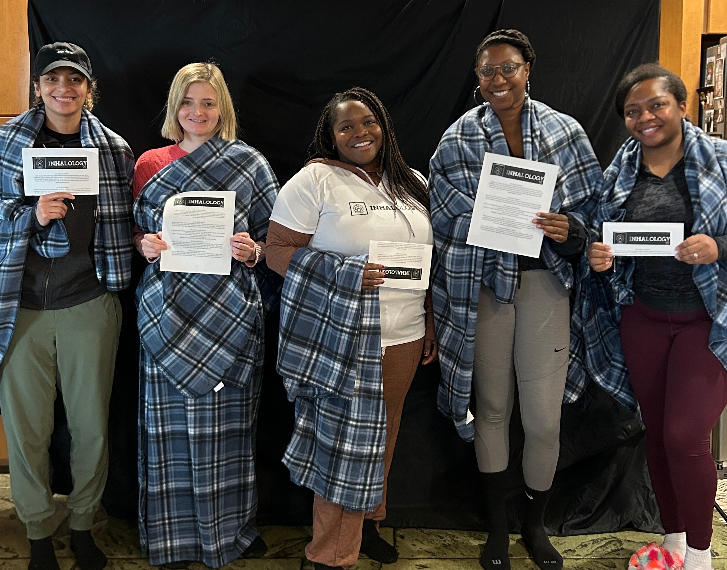 Five women standing together, holding certificates, smiling, with a black background and wooden floor.