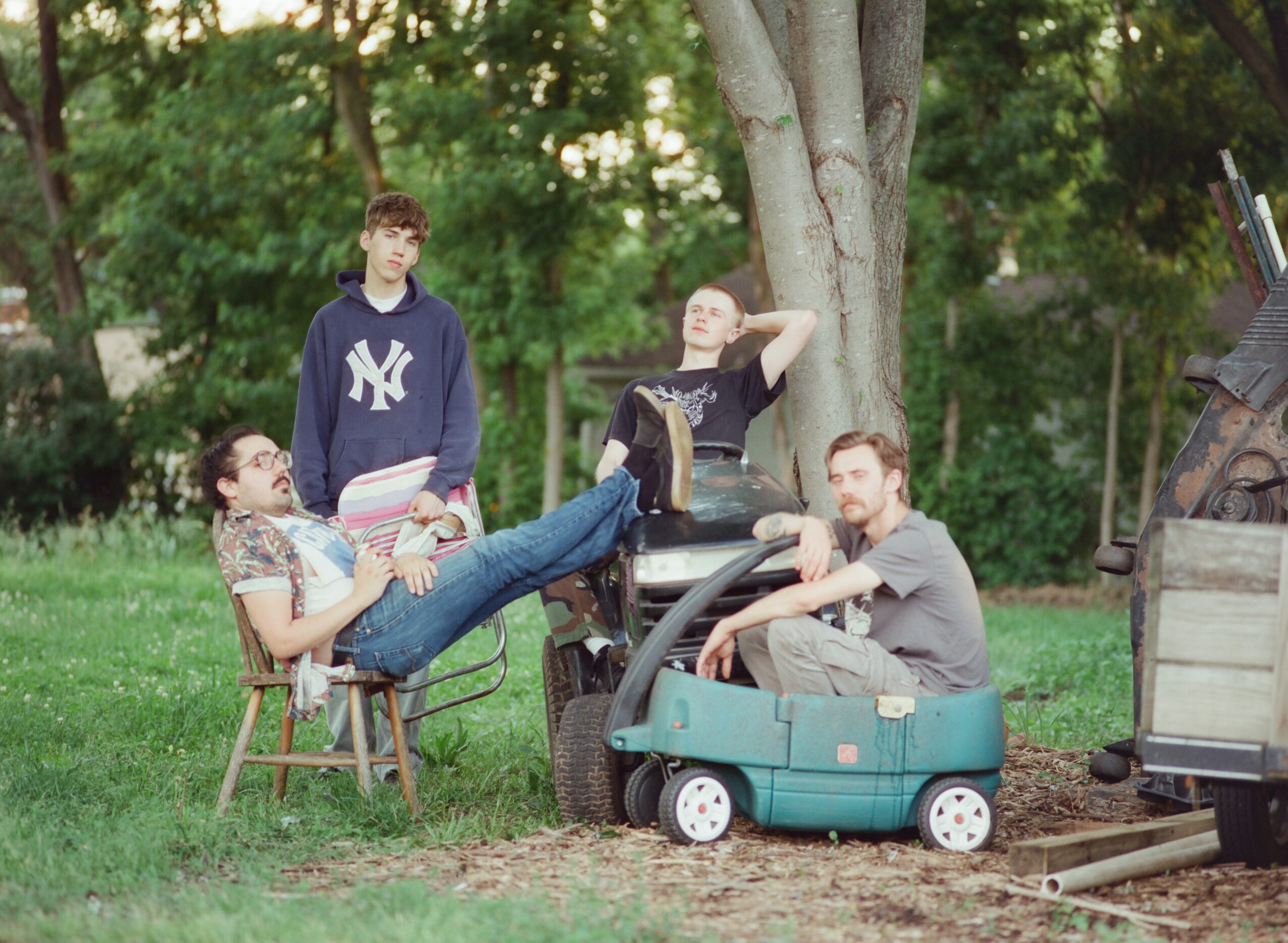 Four people outdoors near a tree, one sitting on a chair, others standing or sitting on a toy car, in a park.