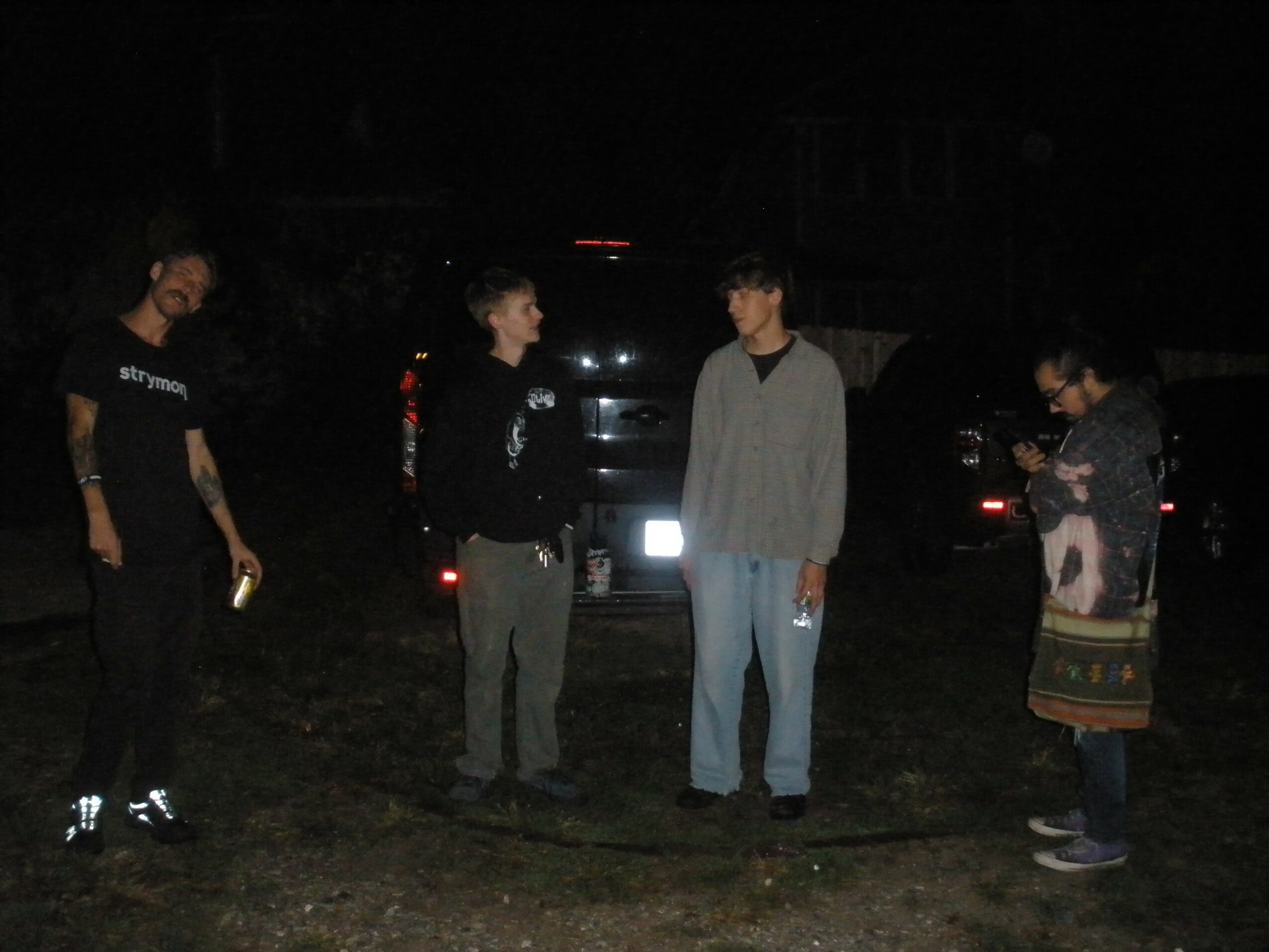 Four people standing outdoors at night, with cars in the background, illuminated by a light source, some holding objects.