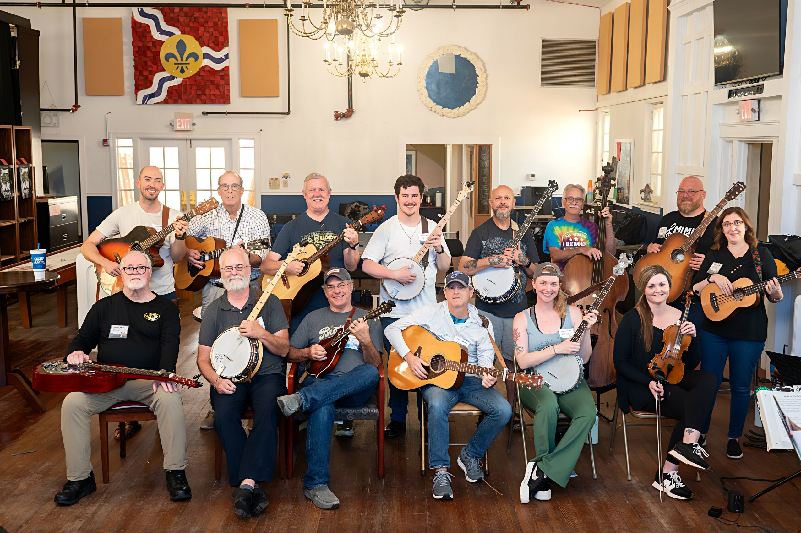 Group of 14 people playing guitars and banjos in a room with artwork and a chandelier, smiling and posing for the photo.